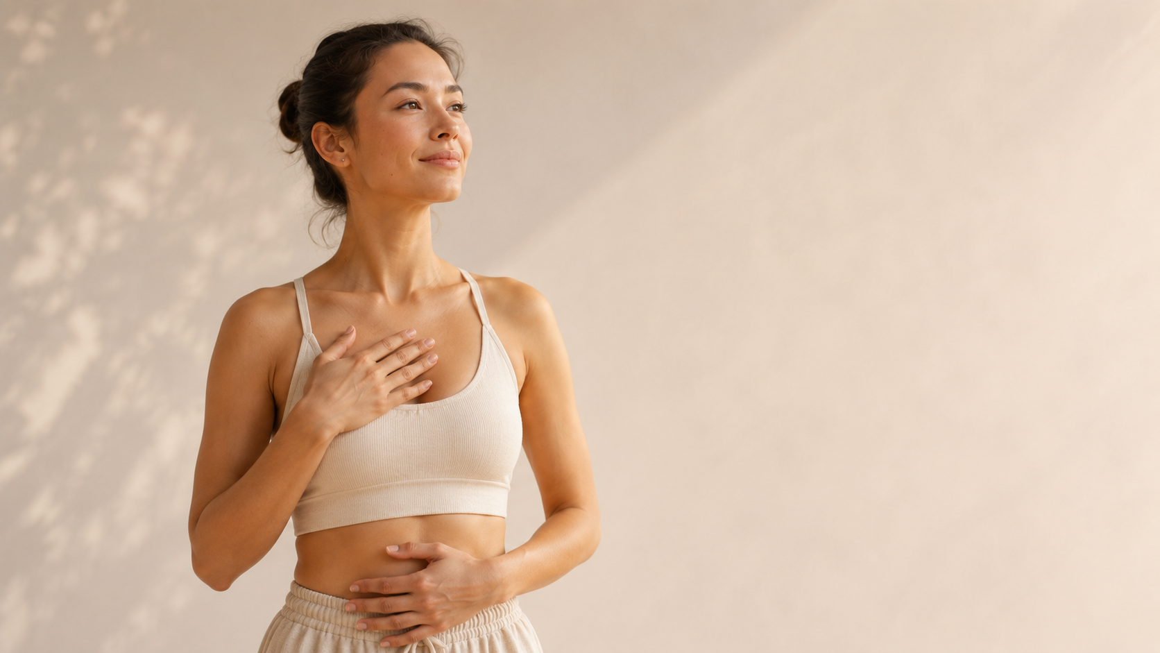 A young woman standing with her hand on her chest and stomach, looking serene and calm, wearing a beige crop top and matching pants against a neutral background.