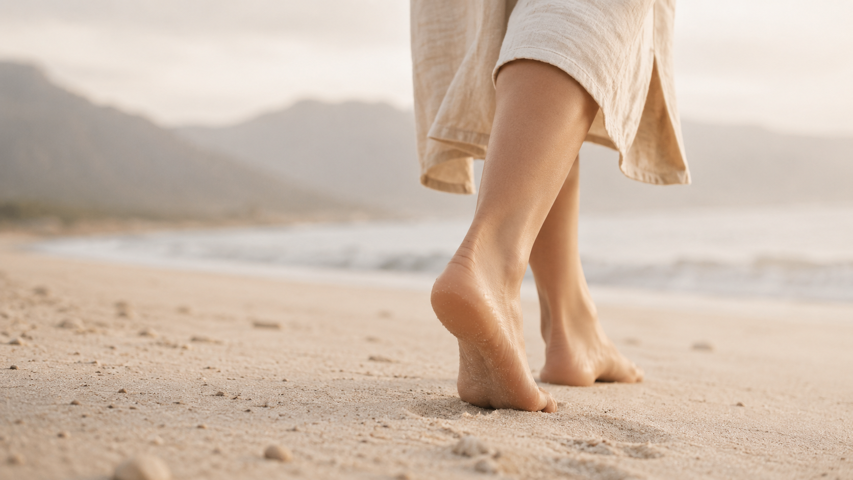 Close-up of a person's feet walking on sandy beach with a towel or wrap around lower legs and ocean in the background.