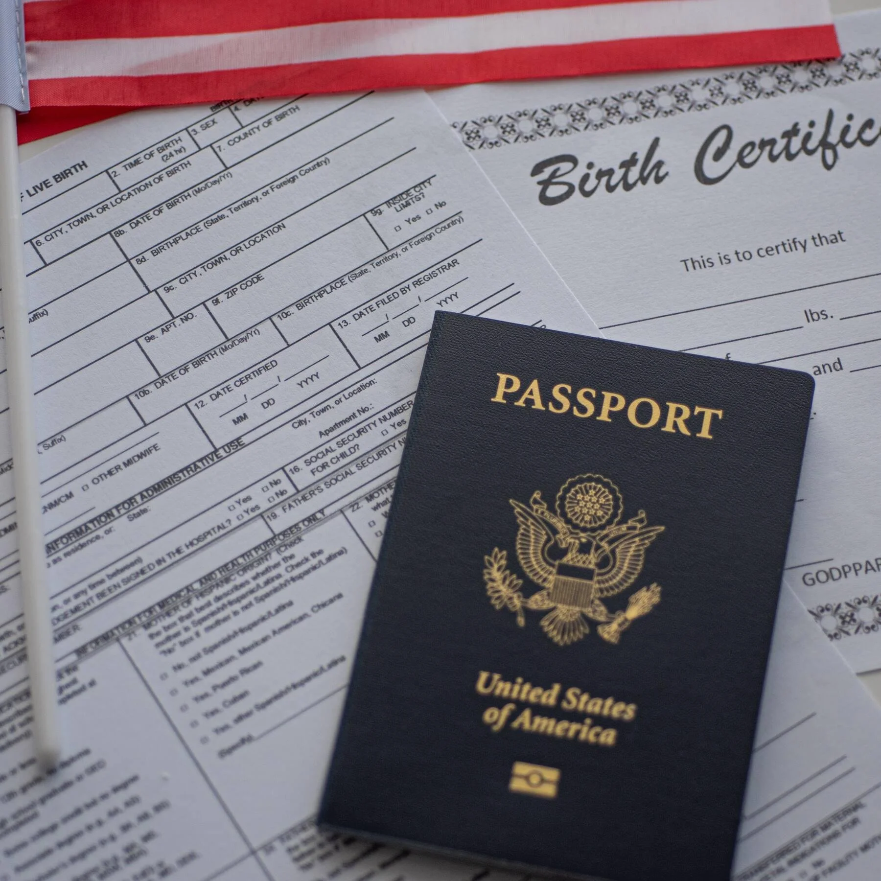 U.S. birth certificate document with an American flag at the top and a black U.S. passport with a gold emblem and text on top of the document.