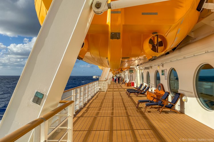 On the deck of a cruise ship, with a yellow lifeboat overhead, blue deck chairs, and ocean in the background.