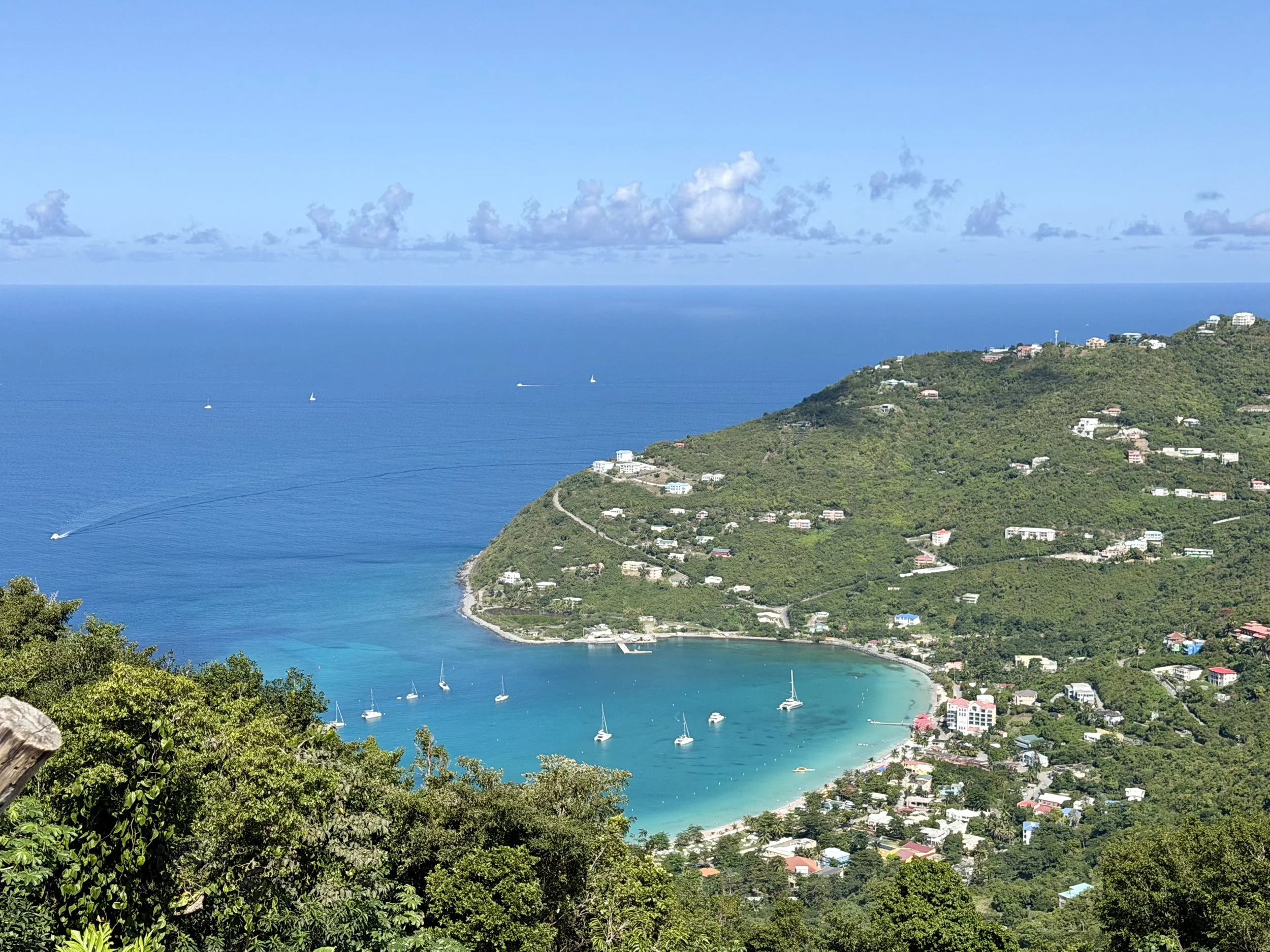 A view of a coastal area with a turquoise bay, sailboats, and green hills dotted with houses and buildings under a blue sky with scattered clouds.