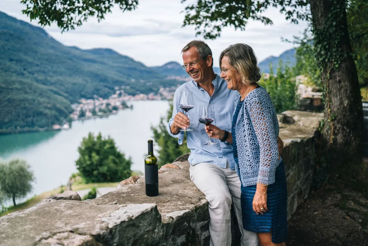 A middle-aged couple enjoying red wine outdoors by a scenic lake with mountains in the background. They are smiling and clinking glasses while standing near a stone wall, with a bottle of wine on the wall.