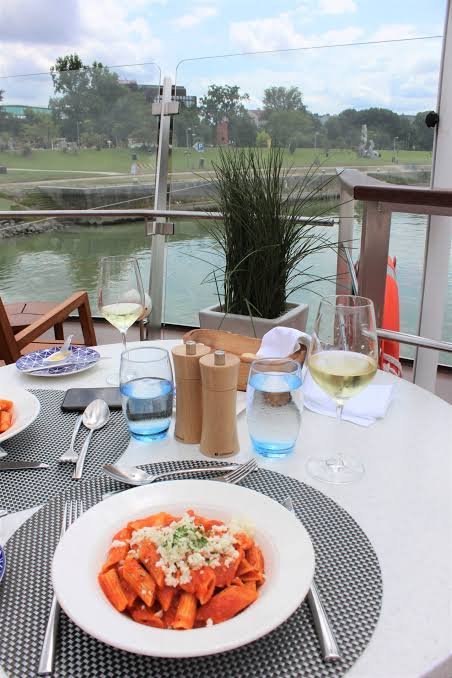 A dining table with pasta, two glasses of white wine, glasses of water, and tableware, set on a balcony overlooking a river and greenery.
