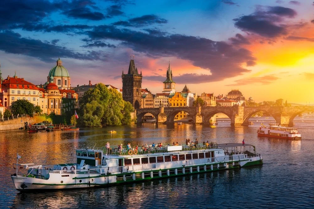 View of Prague with boats on Vltava River during sunset, historic buildings, bridges, and a colorful sky.