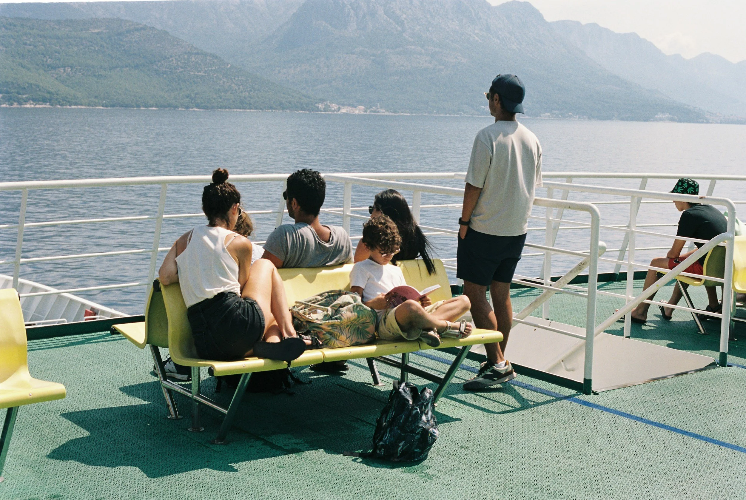 People sitting and standing on the deck of a boat traveling on a large body of water. Mountains are visible in the background.