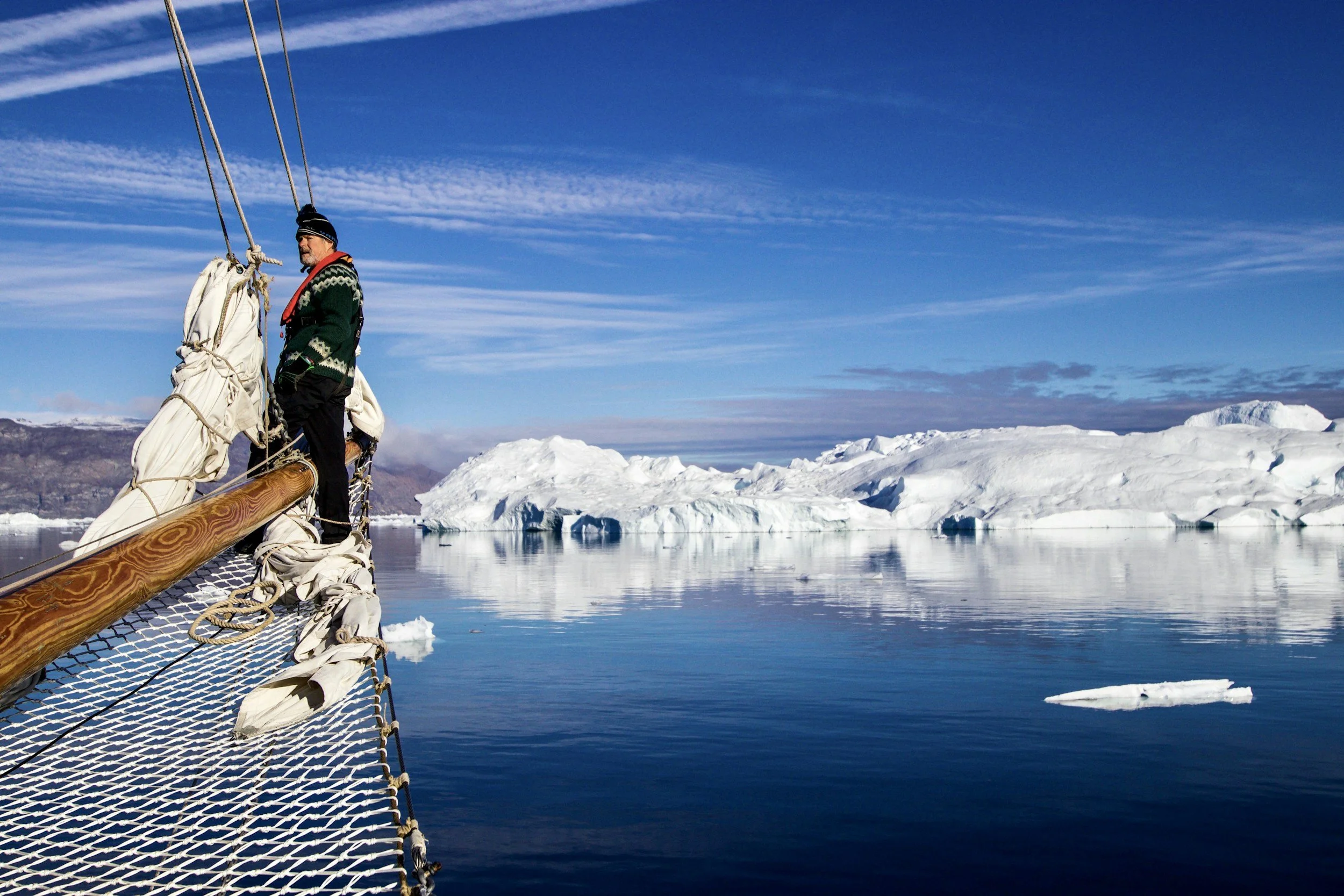 A person wearing a green sweater, black pants, and a beanie hat standing on the bow of a sailboat in icy waters with icebergs in the background, under a blue sky with some clouds.
