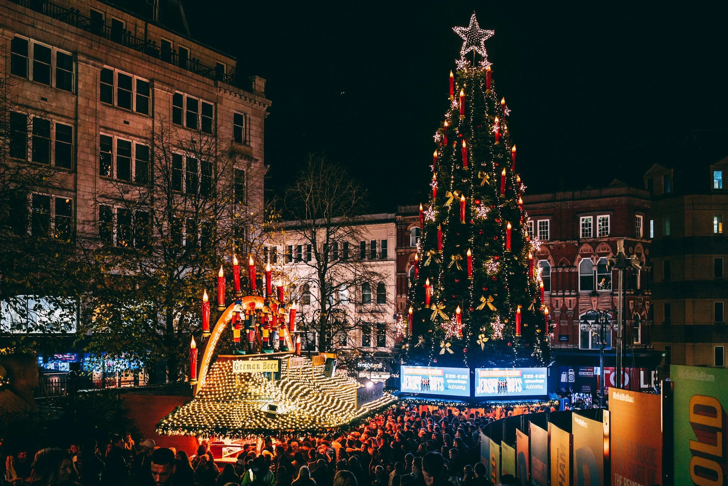 A large decorated Christmas tree with lights, ornaments, and a star topper at night in an urban plaza, surrounded by a crowd of people and illuminated booths, with buildings visible in the background.