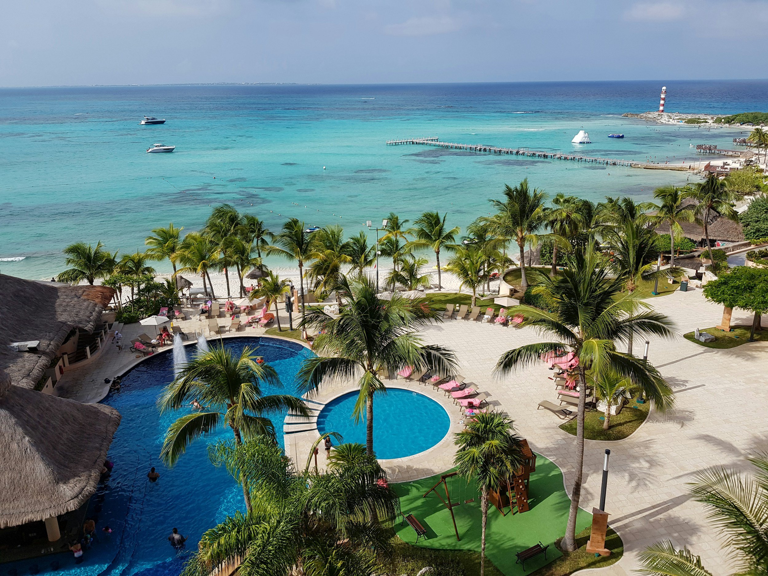 A tropical resort with a swimming pool surrounded by palm trees, lounge areas, and a view of the ocean with boats and a lighthouse in the distance.