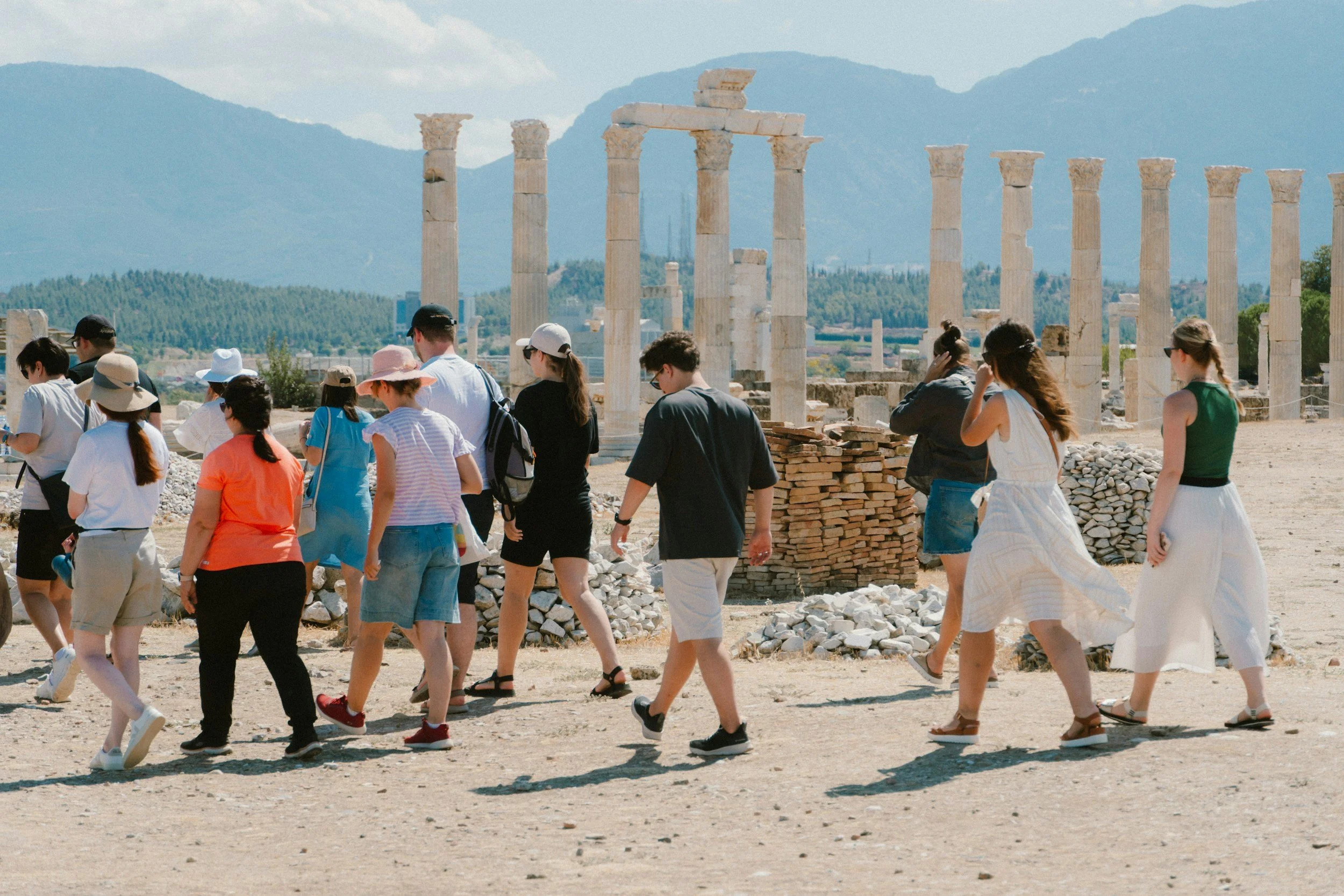 A group of tourists walking among ancient ruins with tall stone columns, mountain scenery in the background, and a clear sky.