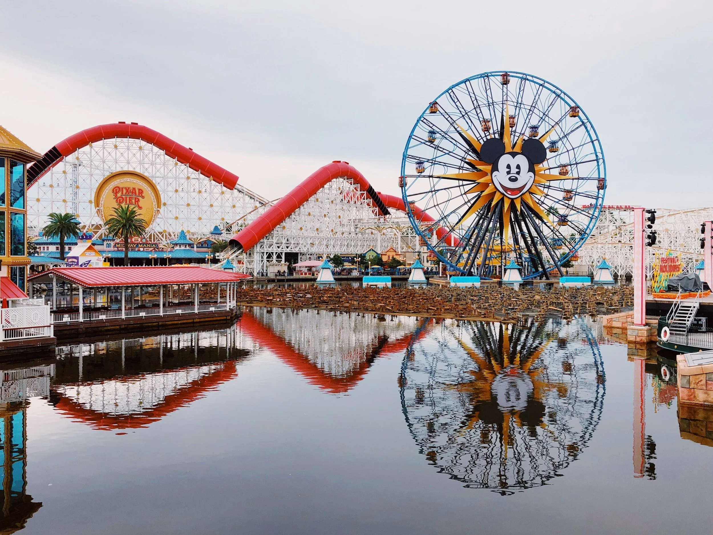 A view of the Pixar Pier at a Disney amusement park with a large Mickey Mouse Ferris wheel and a roller coaster, reflected in the water below.