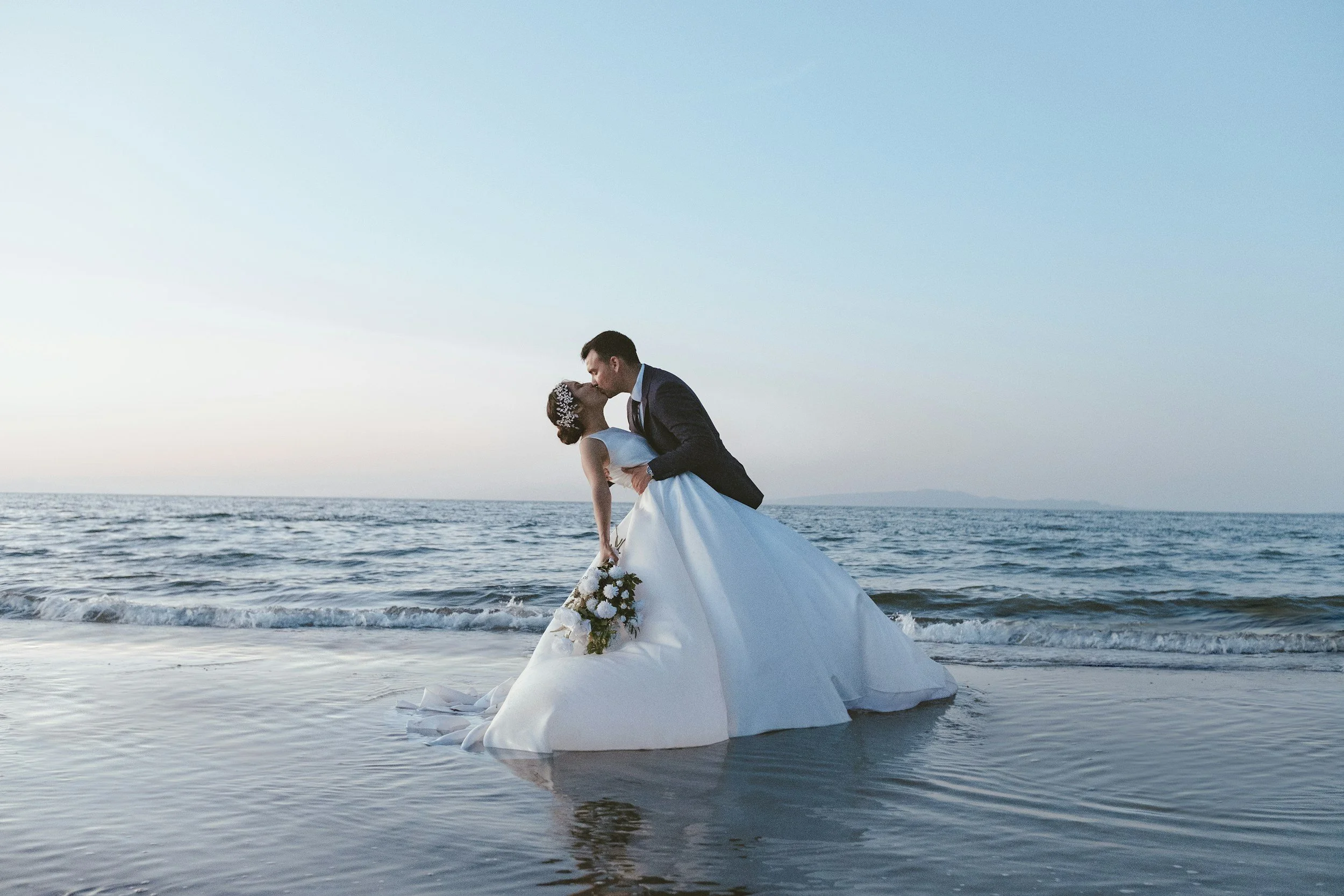 A bride and groom sharing a kiss on a boat in the ocean at sunset, with the bride holding a bouquet of white flowers.
