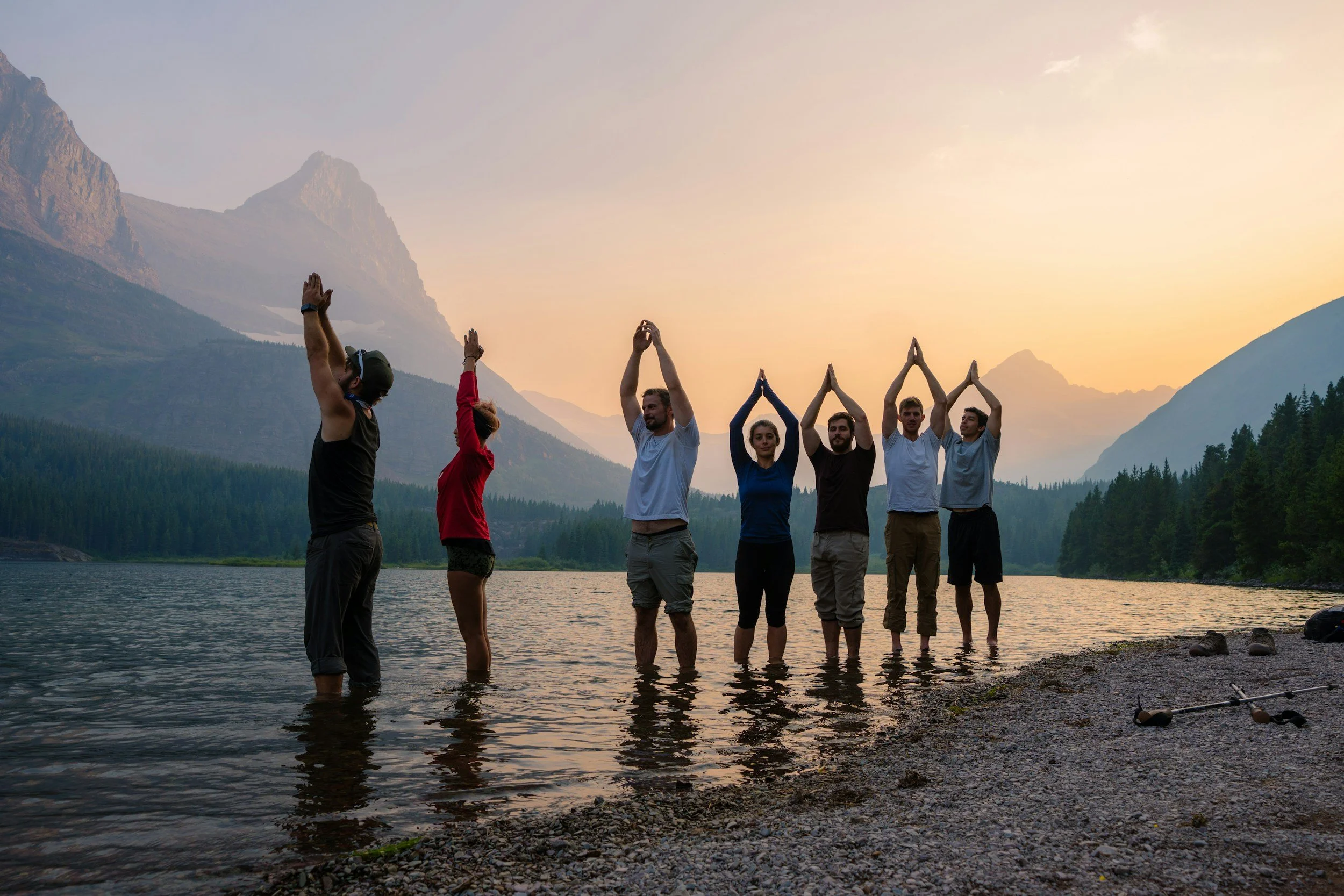 A group of people standing in a line in shallow water at the edge of a lake, performing yoga poses with their hands raised above their heads in front of a mountain landscape during sunset.