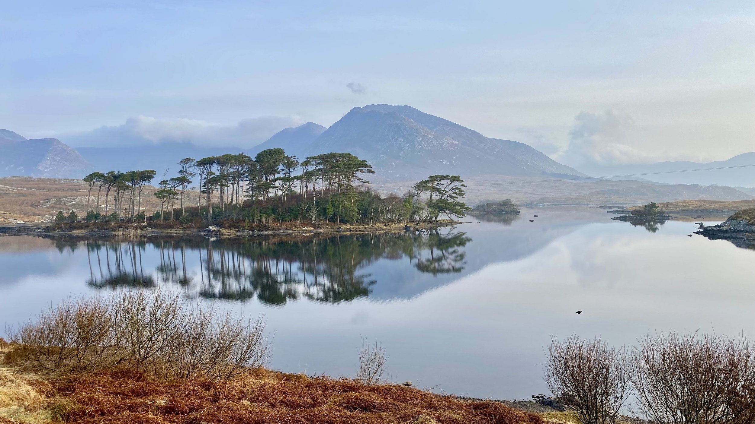 Lake with calm reflective water, surrounded by trees and mountains in the distance under a partly cloudy sky.