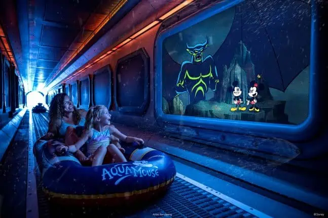 Two young girls sitting in an inflatable boat labeled 'Aqua Mouse' on a dark water ride at an amusement park, watching animated characters on a large water-themed display, including a large fish-like creature and Mickey Mouse and Minnie Mouse.