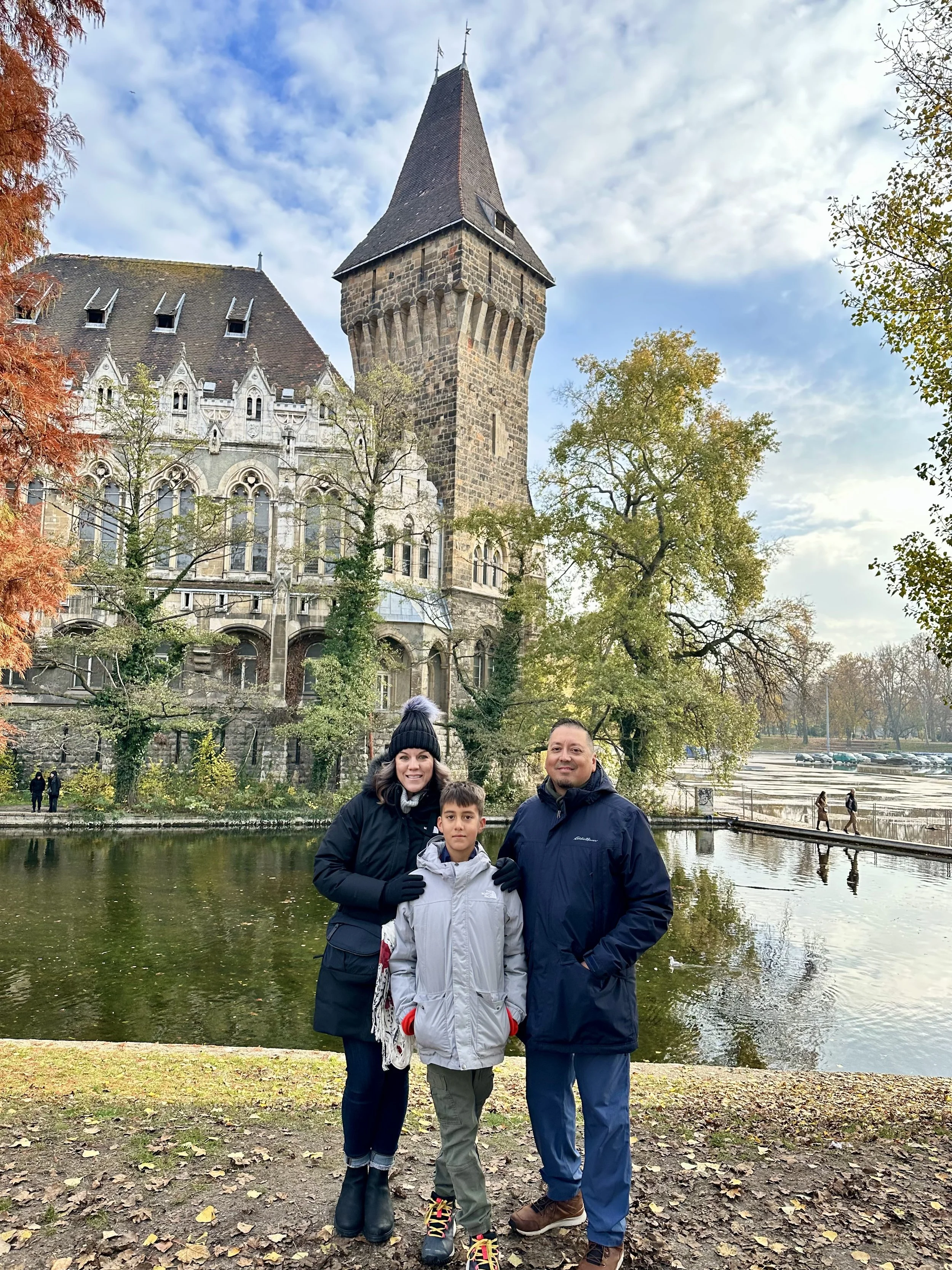A family of three standing near a pond with trees and a historic stone building featuring a tall tower in the background.