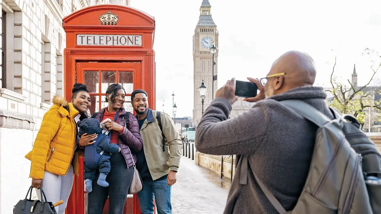 Three happy tourists taking a photo of a family in front of a red telephone booth with Big Ben in the background in London.