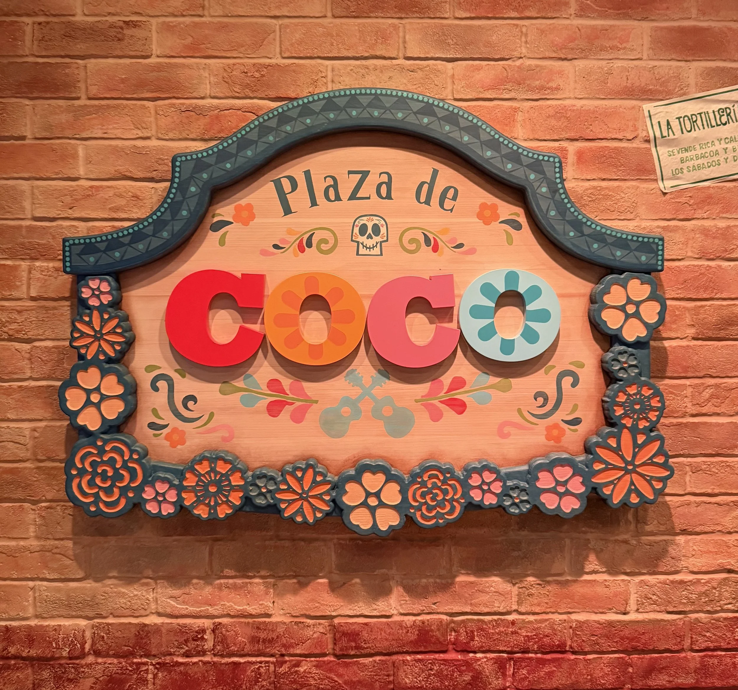 Decorative signboard with the words "Plaza de" and colorful floral and skull motifs, mounted on a red brick wall.