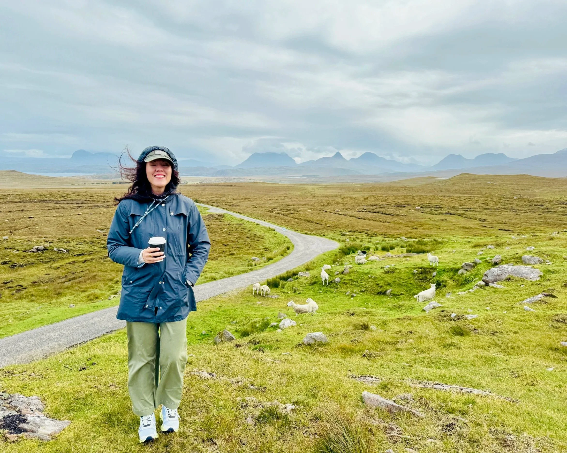 A woman wearing a blue rain jacket, khaki pants, and a beige cap is holding a cup, standing on a grassy path in an open field with sheep grazing nearby. In the background, there are mountains and a cloudy sky.