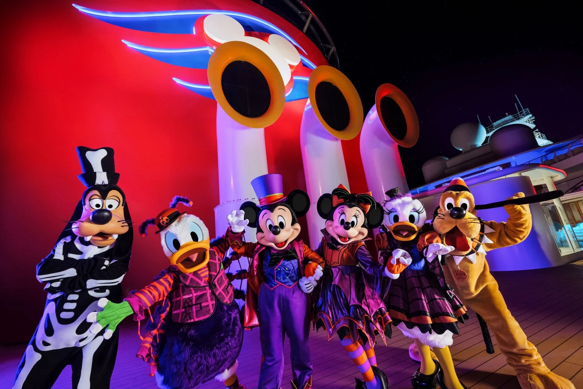 Disney characters dressed in Halloween costumes posing in front of a colorful Mickey Mouse shaped structure at night.