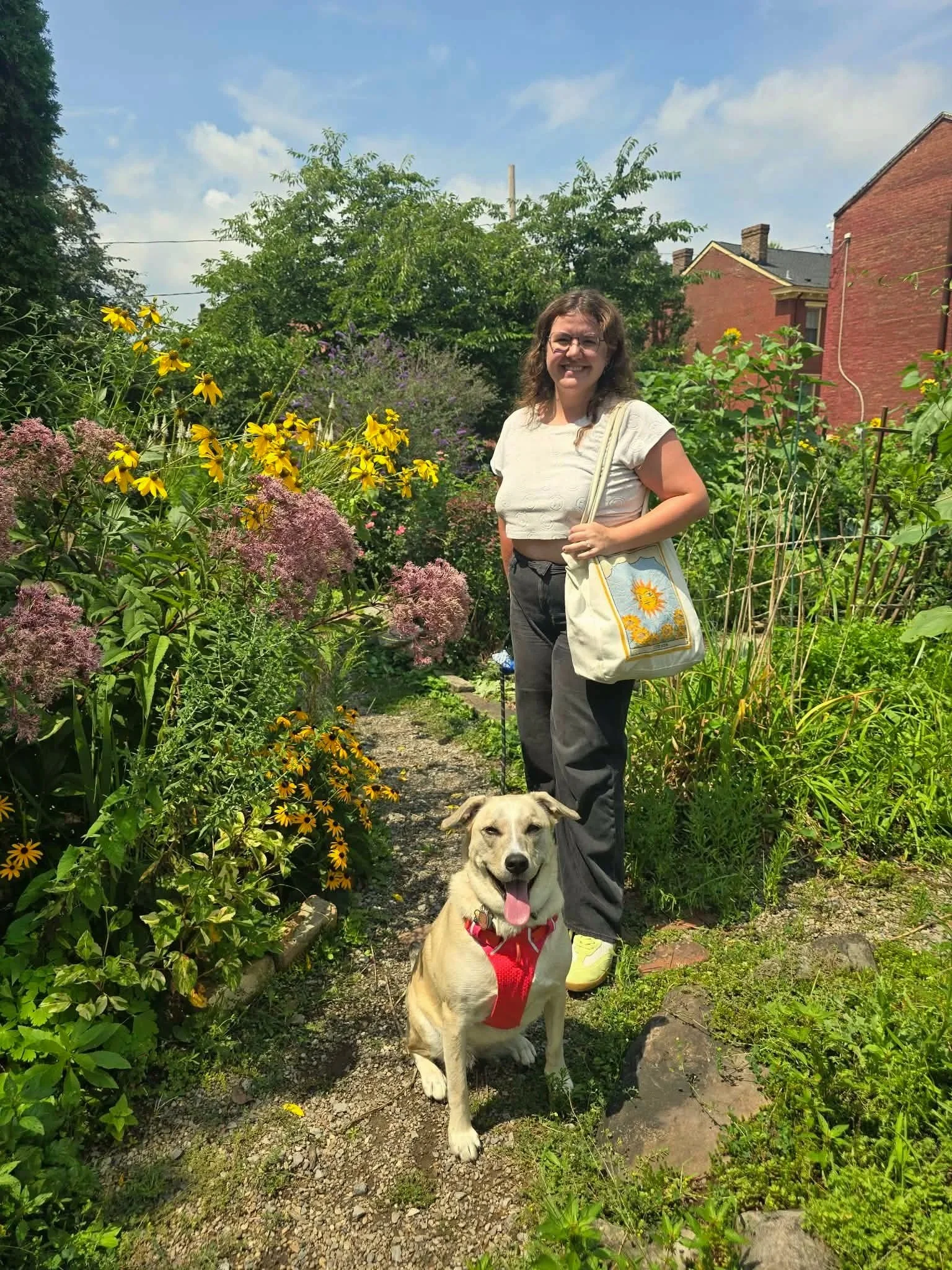 A woman with glasses and curly brown hair standing on a garden path next to a large yellow and white dog with a red bandana, surrounded by various green plants, yellow and purple flowers, and a red brick house in the background under a partly cloudy sky.