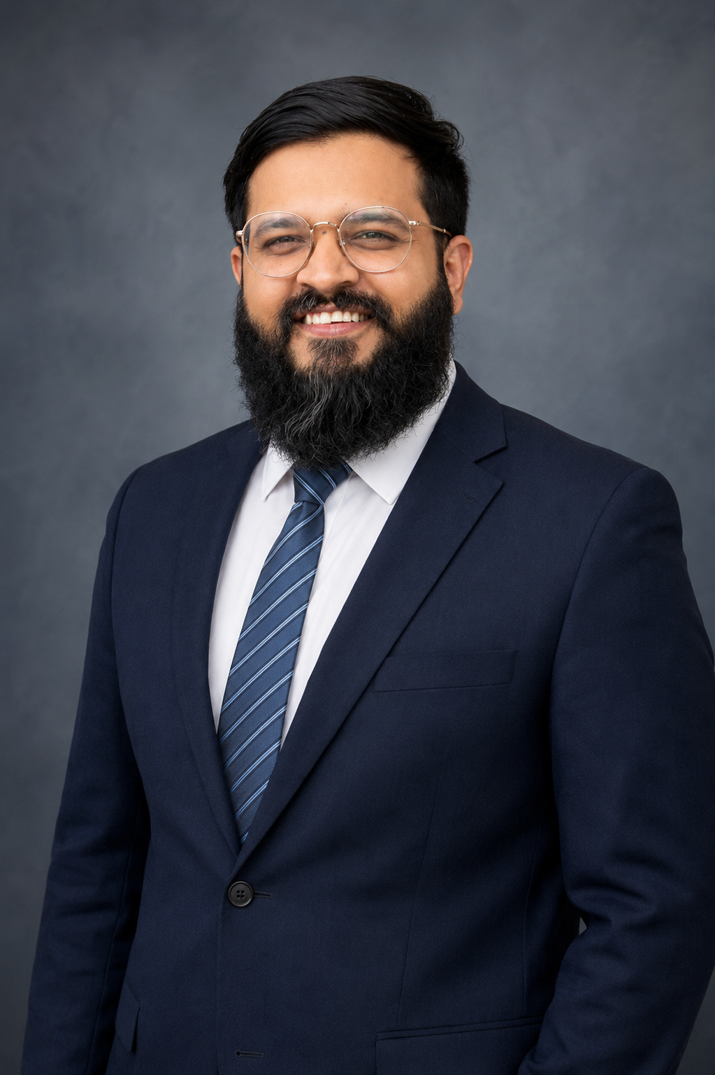 A man with glasses and a beard wearing a dark blue suit, white shirt, and blue striped tie, smiling against a gray background.