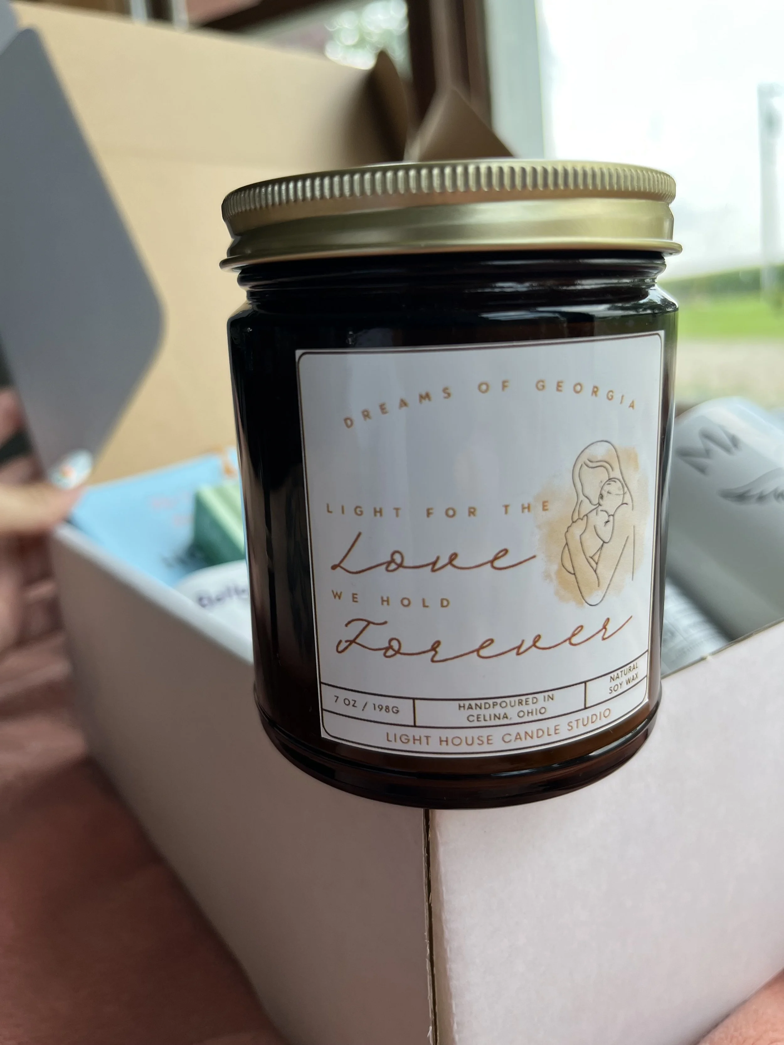 Close-up of a candle jar with a label that features an illustration of a mother holding a baby, and text that reads 'Dreams of Georgia, Light for the Love, We Hold Forever, Handpoured in Celina, Ohio, Light House Candle Studio.' The background shows a window with sunlight.
