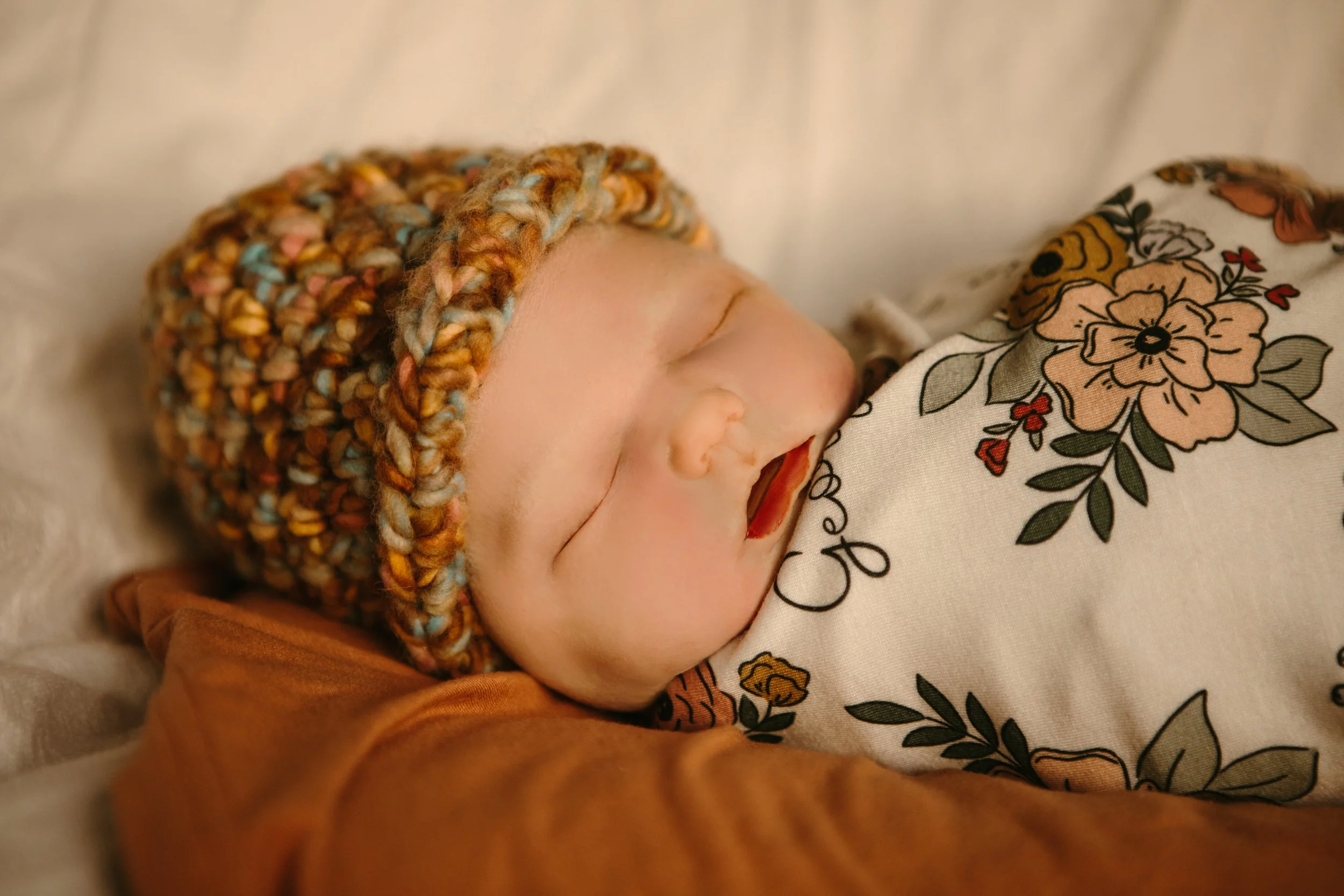 Close-up of a baby born sleeping wearing a and multicolored knit hat, swaddled in floral patterned cloth.