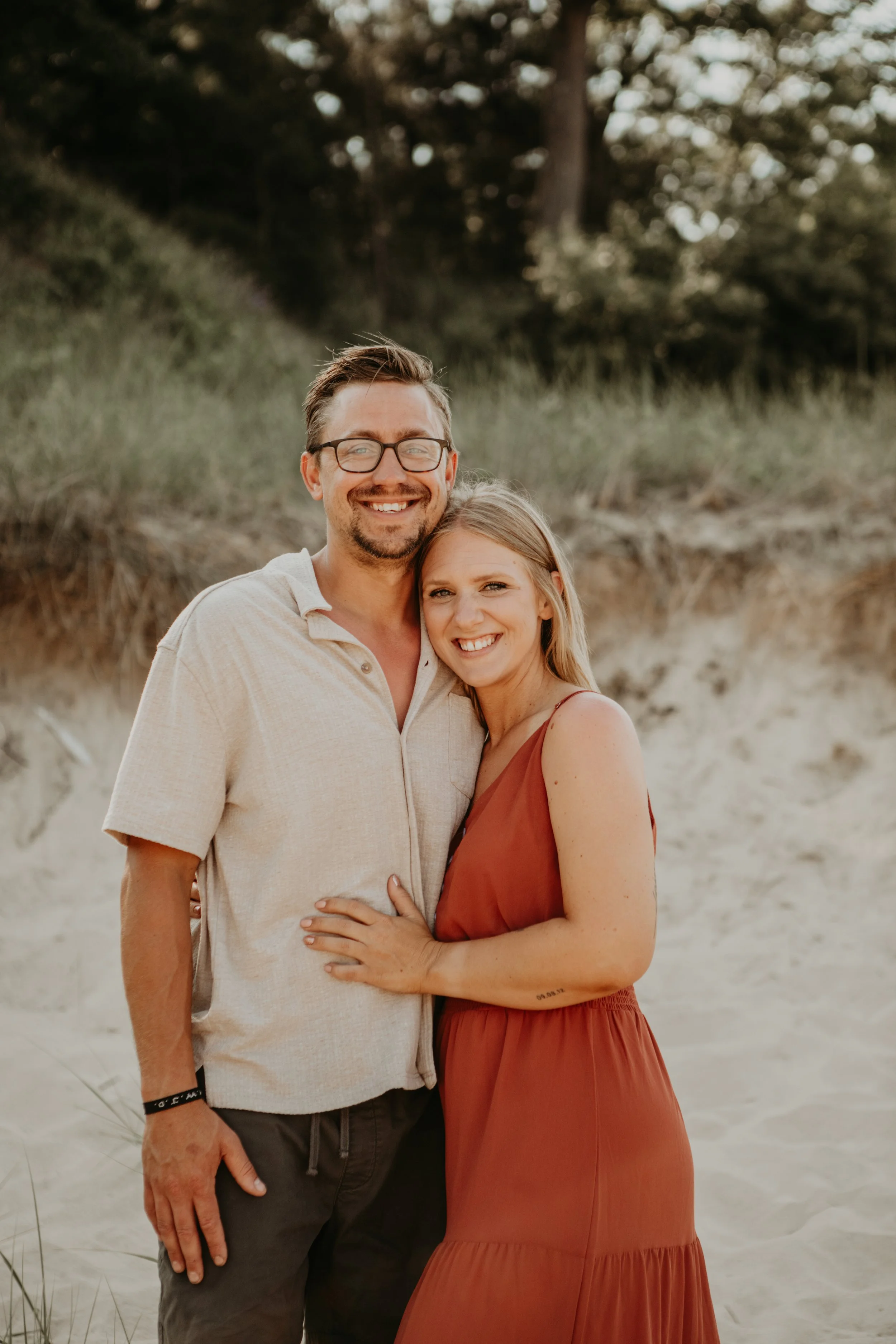 A smiling man with glasses and a beard, and a woman with blonde hair, both standing on a beach with sandy dunes and tall grass in the background, enjoying a sunny day.