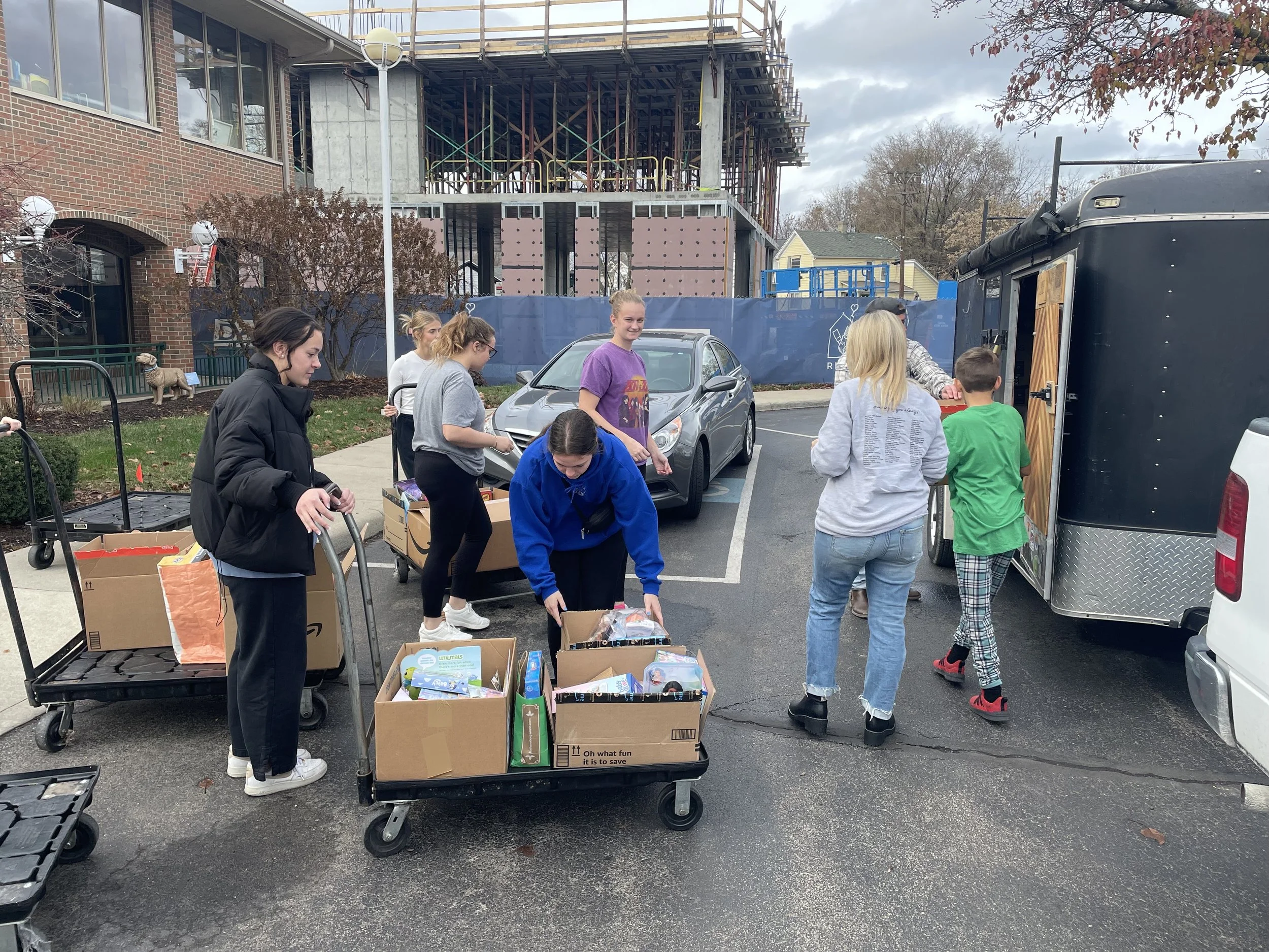 People gather outdoors near a food truck and boxes of snacks or supplies, with a building under construction in the background.