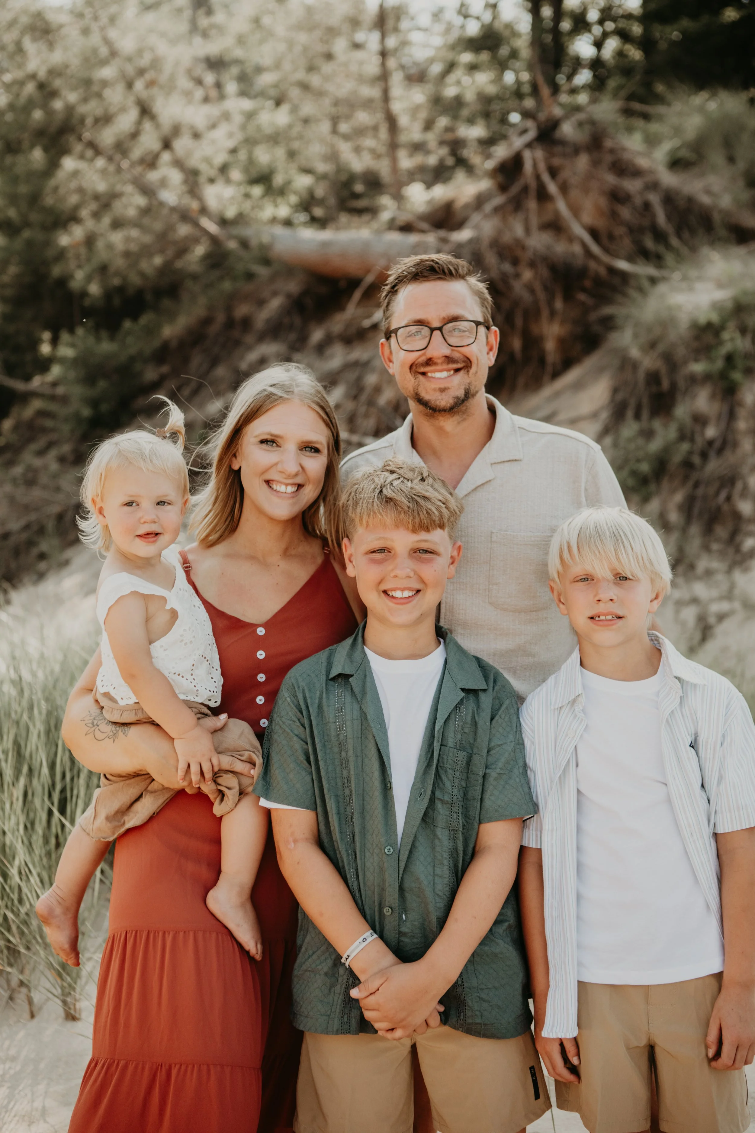 Family of five smiling at the beach, standing in front of trees and sand.
