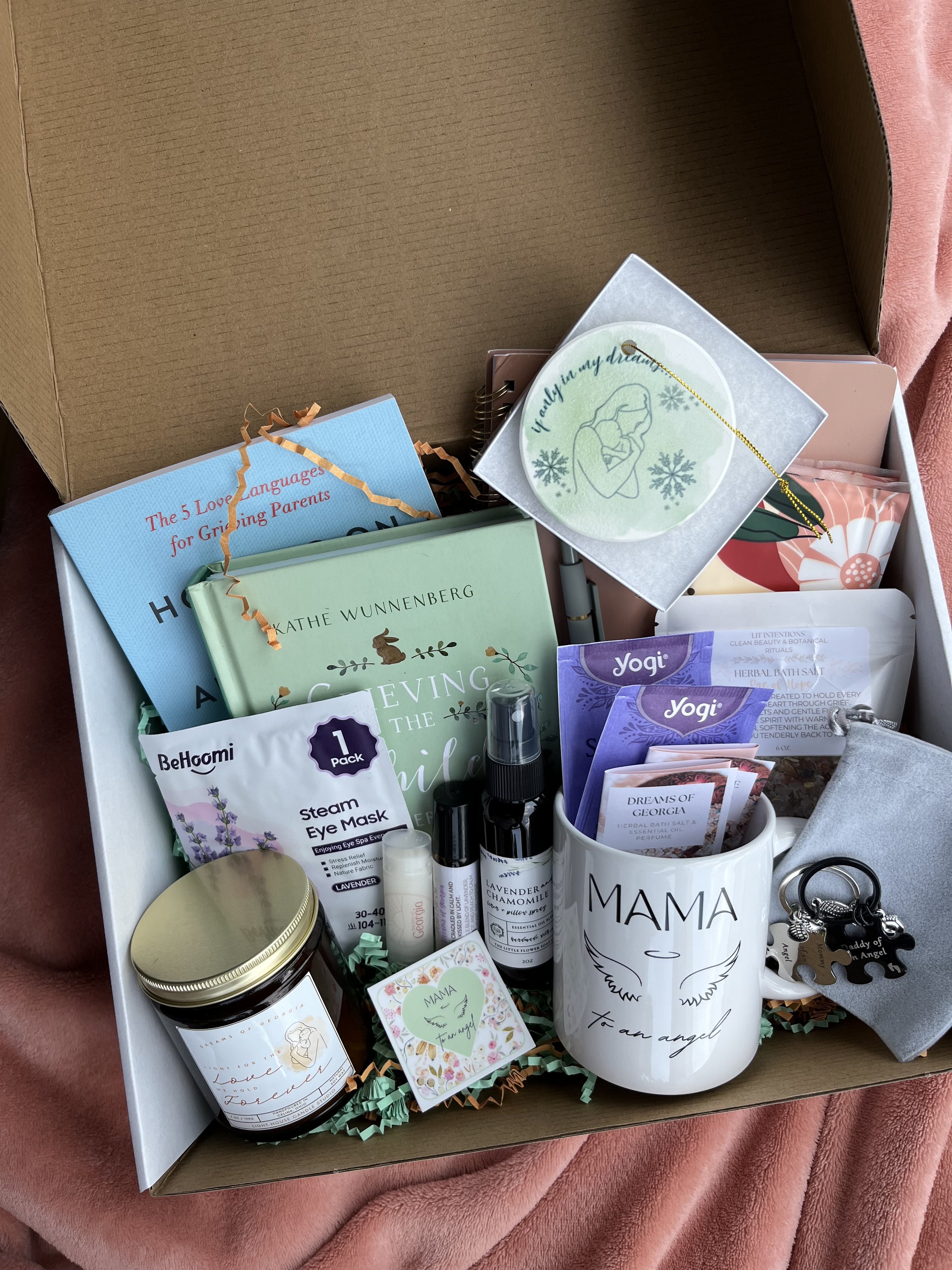 A Rae of Hope box containing various self-care and spiritual items, including candles, essential oil sprays, skincare masks, a mug, and assorted packets and books, on a pink blanket.