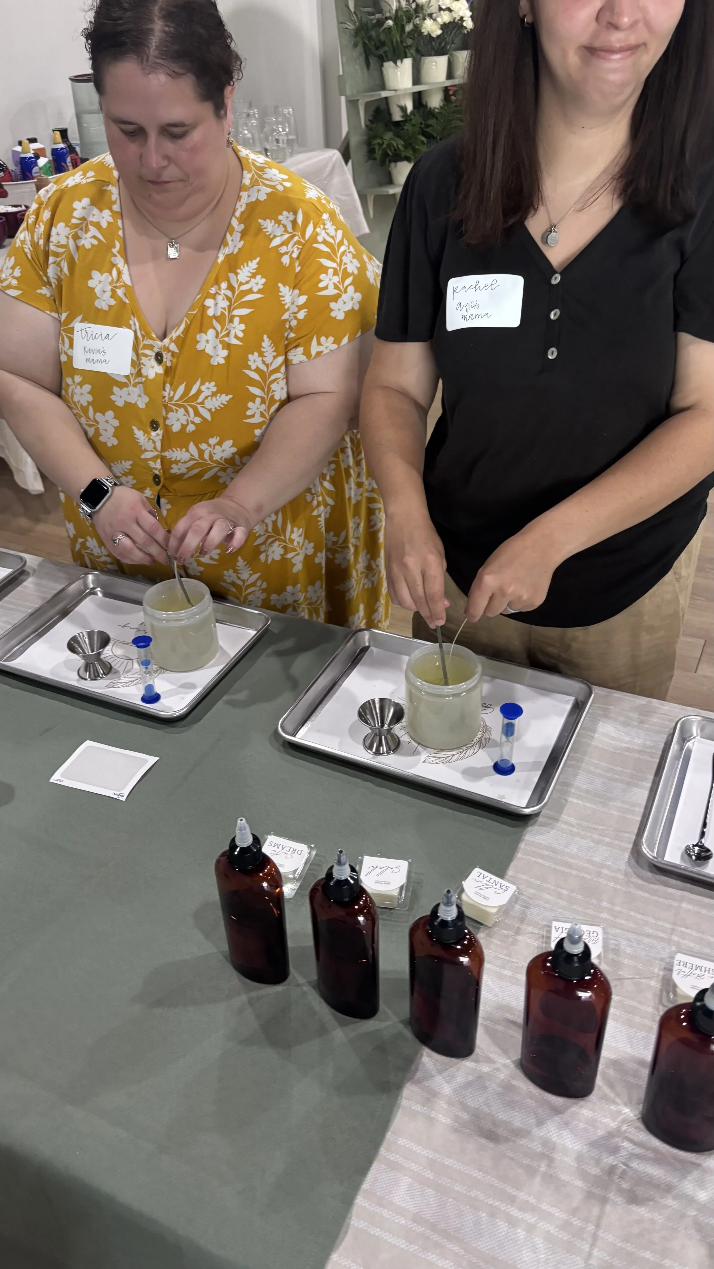 Two women standing at a table, engaging in a candle-making activity, with various supplies and bottles of wax in front of them. This was taken at a Gathering of Hope group meeting.
