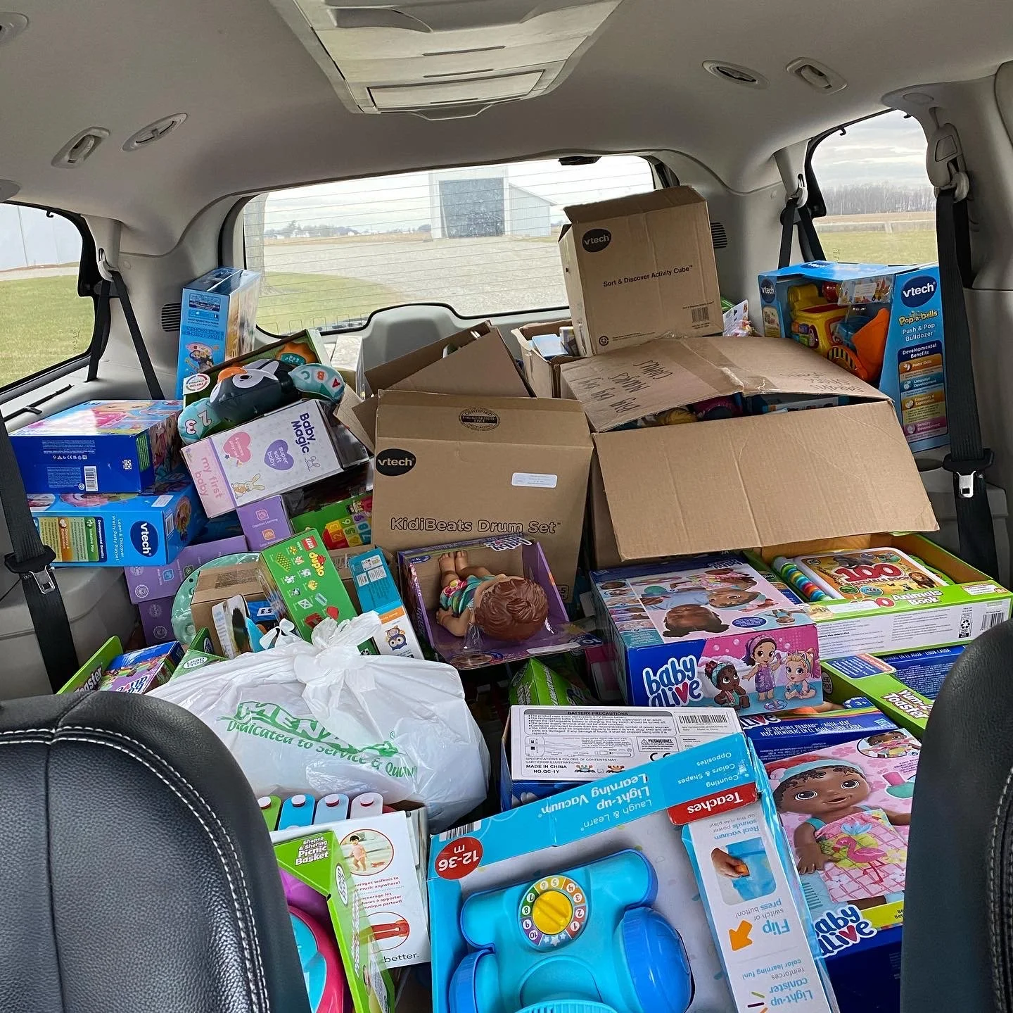 Packed car trunk filled with various toys and boxes, including doll sets, educational toys, and activity cubes, with a scenic outdoor view through the rear window.