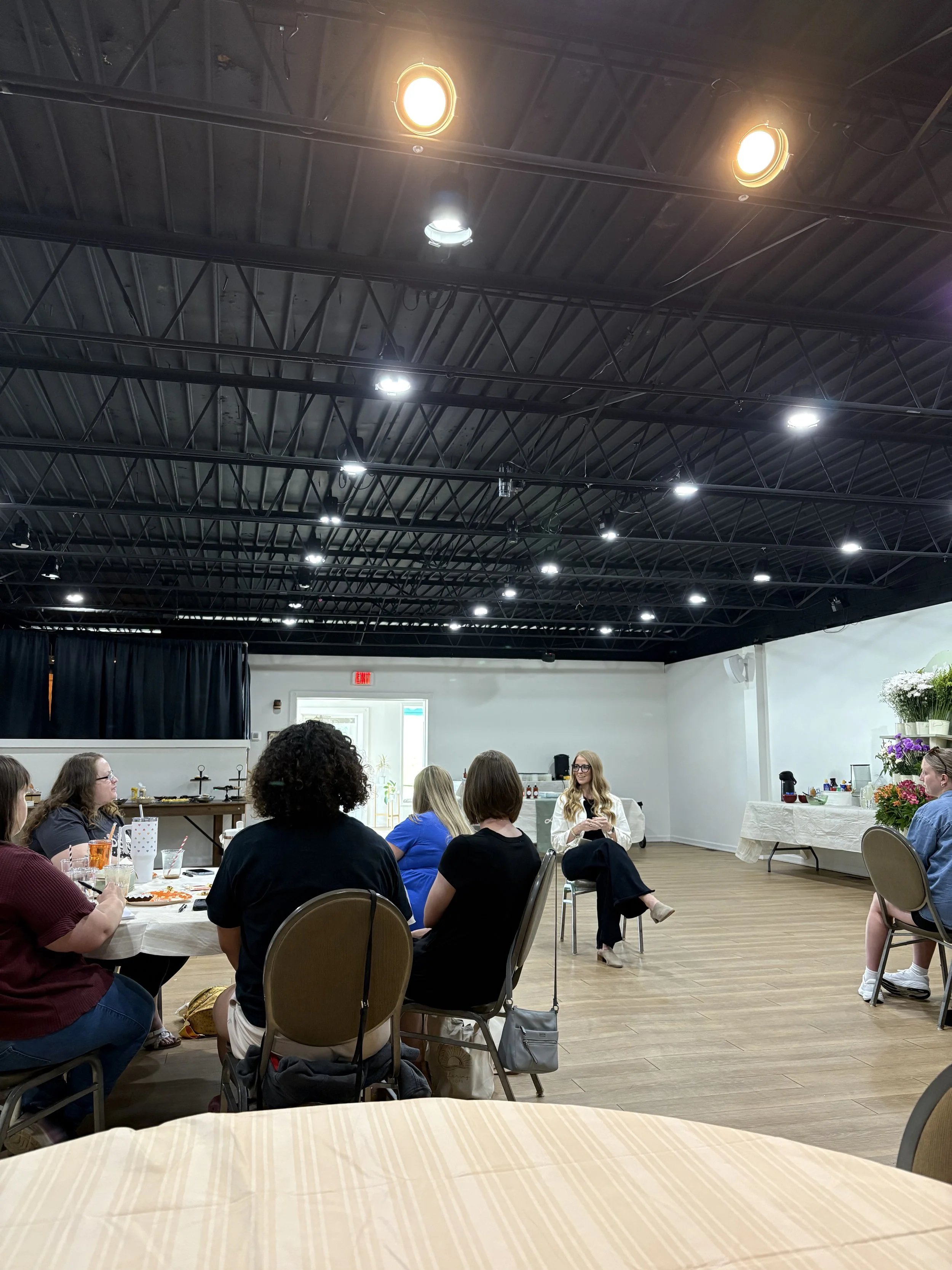 People sitting at tables in a large room with a high ceiling and bright lighting, some engaged in conversation, and a woman standing and speaking at the front. This was taken at a Gathering of Hope group meeting.