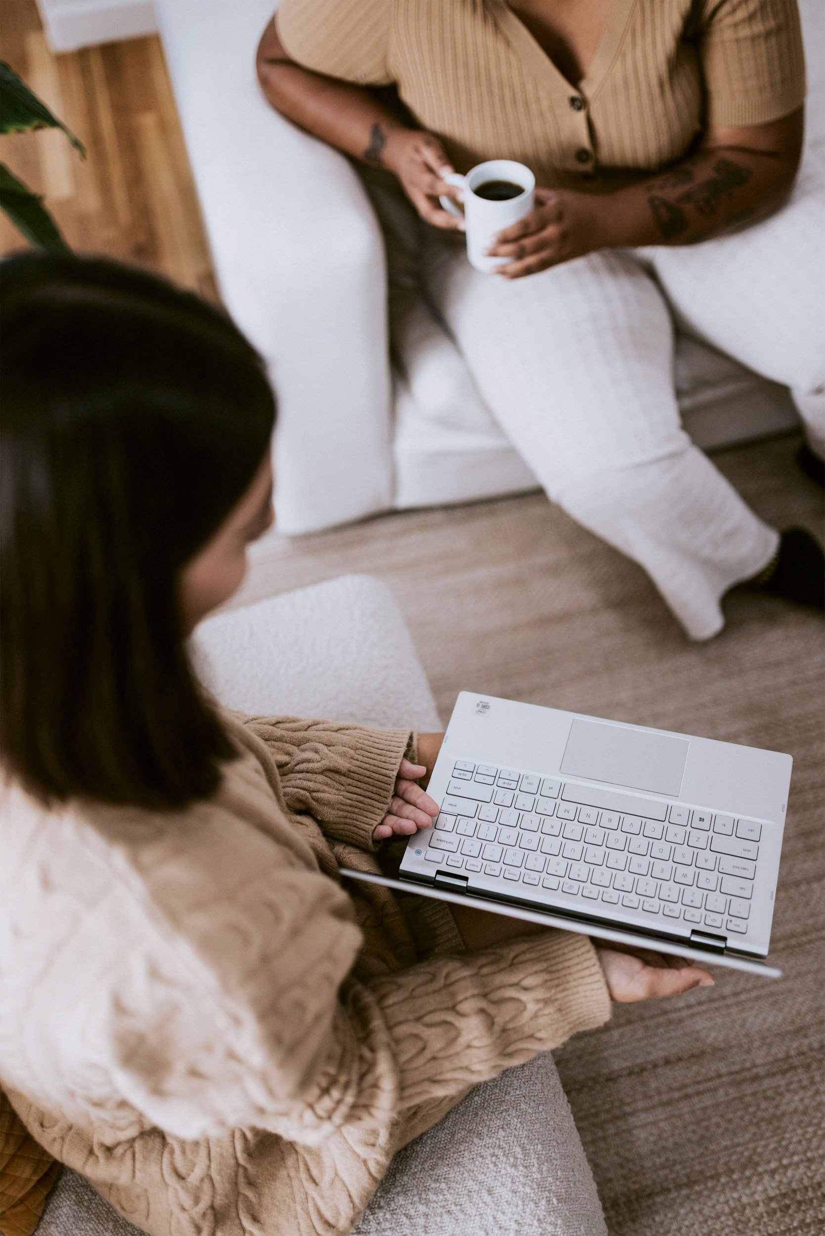 A person sitting on a beige couch holding a white keyboard with their lap resting on a beige blanket. Another person is sitting nearby holding a white cup of coffee, wearing a striped shirt and white pants. A third person is partially visible, holding a white document or notebook.