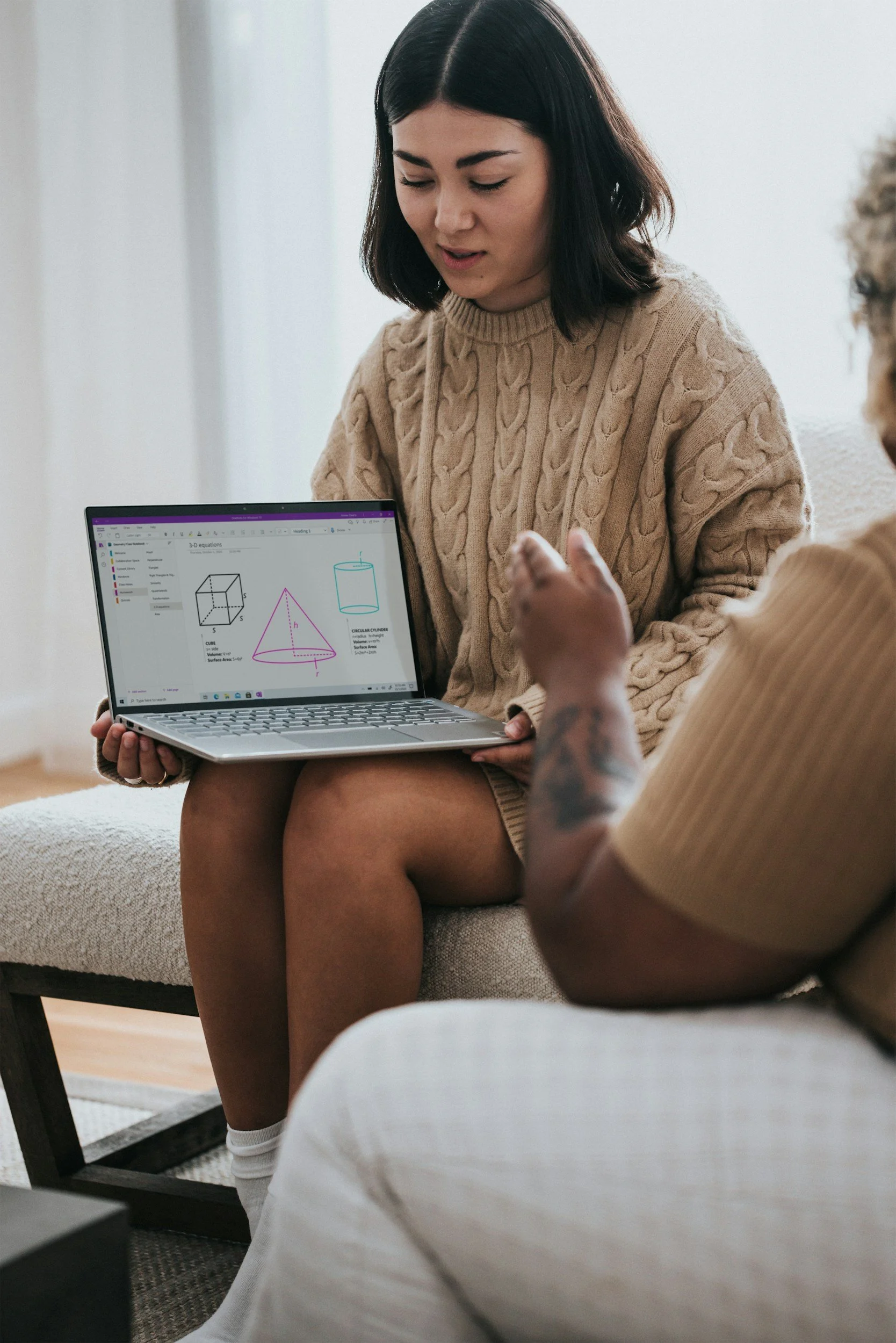 A woman is sitting on a couch, looking at a laptop that displays diagrams of 3D geometric shapes, while another person with a tattoo on their arm faces her.