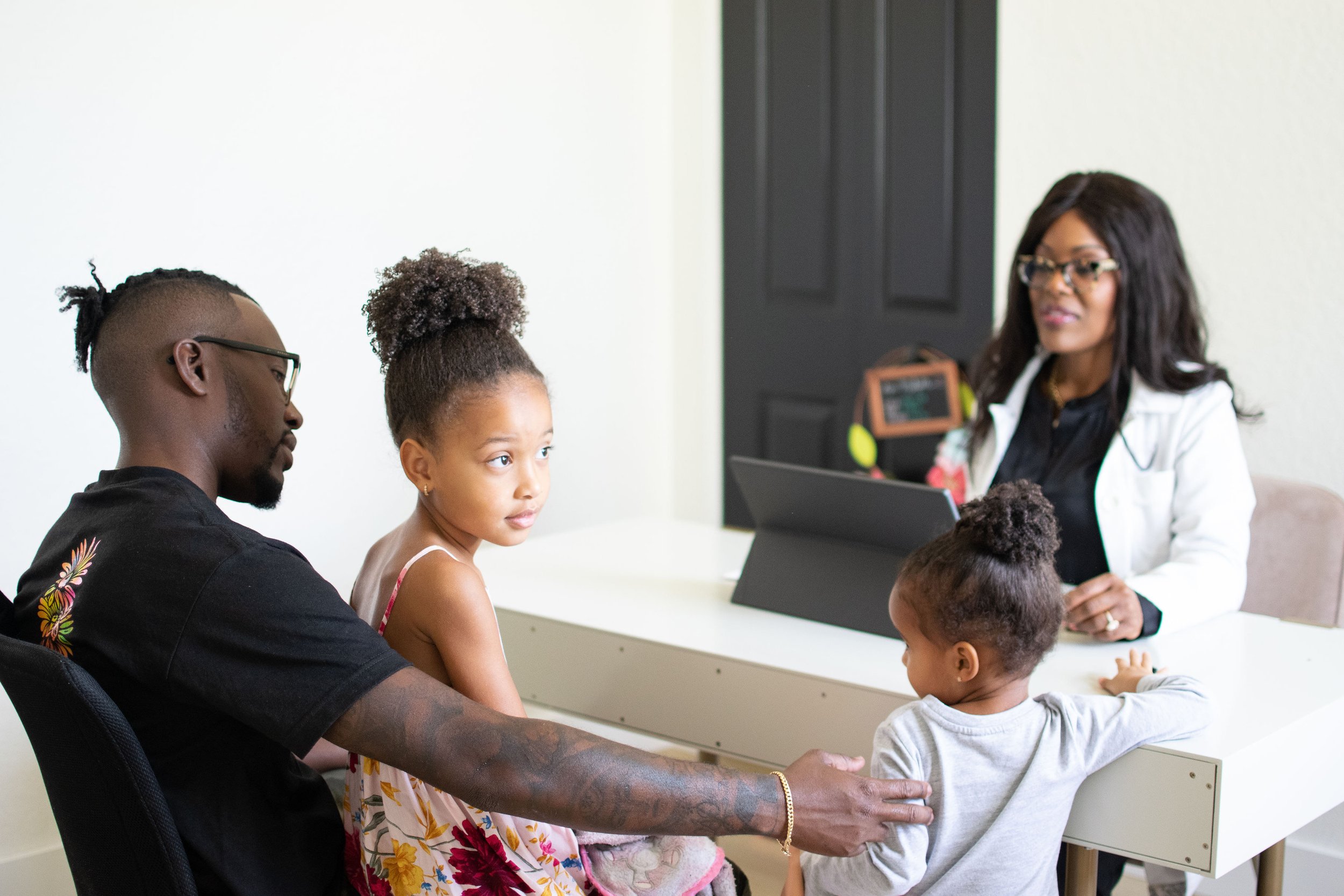 A diverse group of children and a woman with glasses and long dark hair sitting at a white table in a medical or consultation setting, engaging with a healthcare professional, possibly for a check-up or consultation.