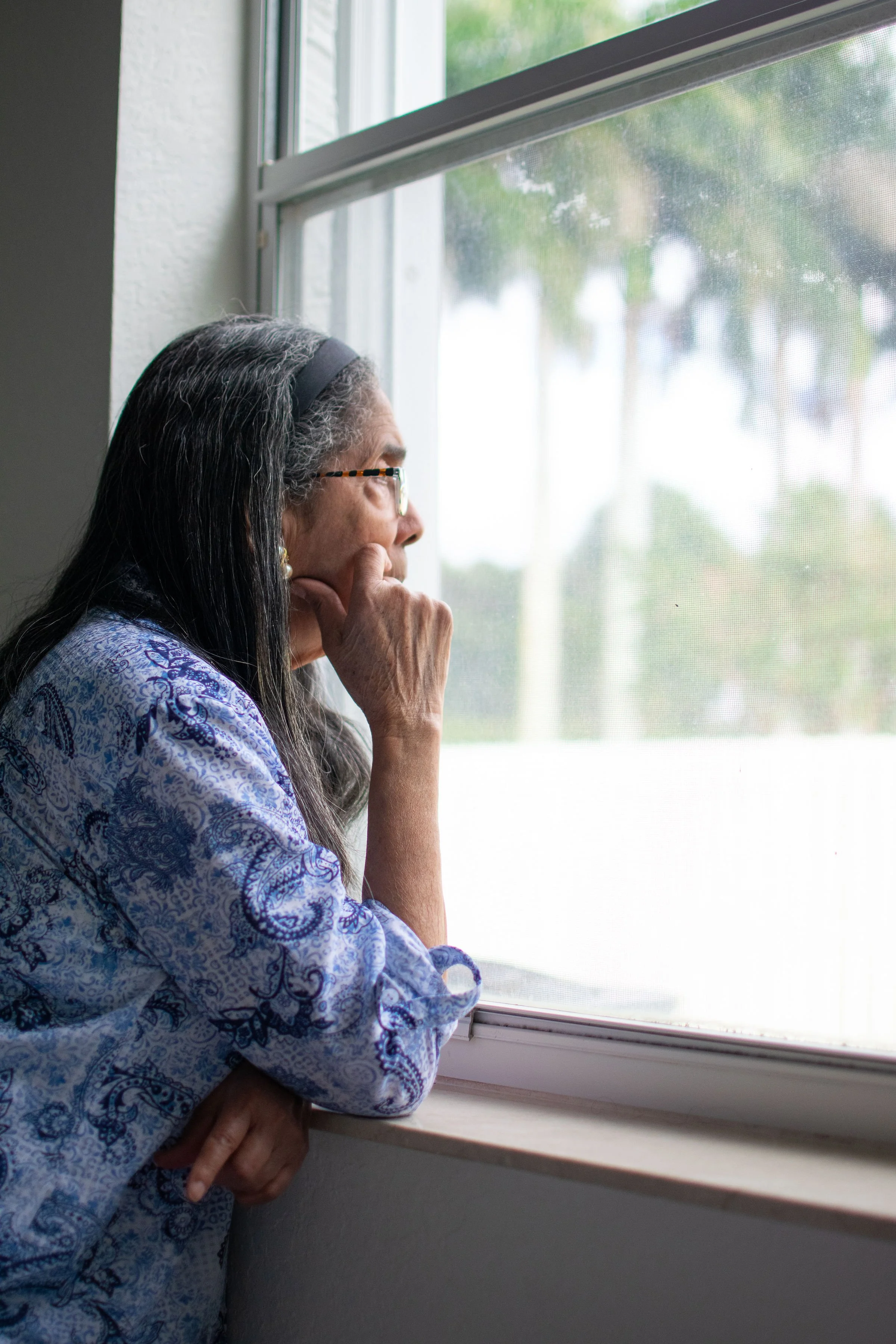 Older woman with long gray hair and glasses, wearing a blue paisley-patterned shirt, looking out a window with a screen, resting her chin on her hand.