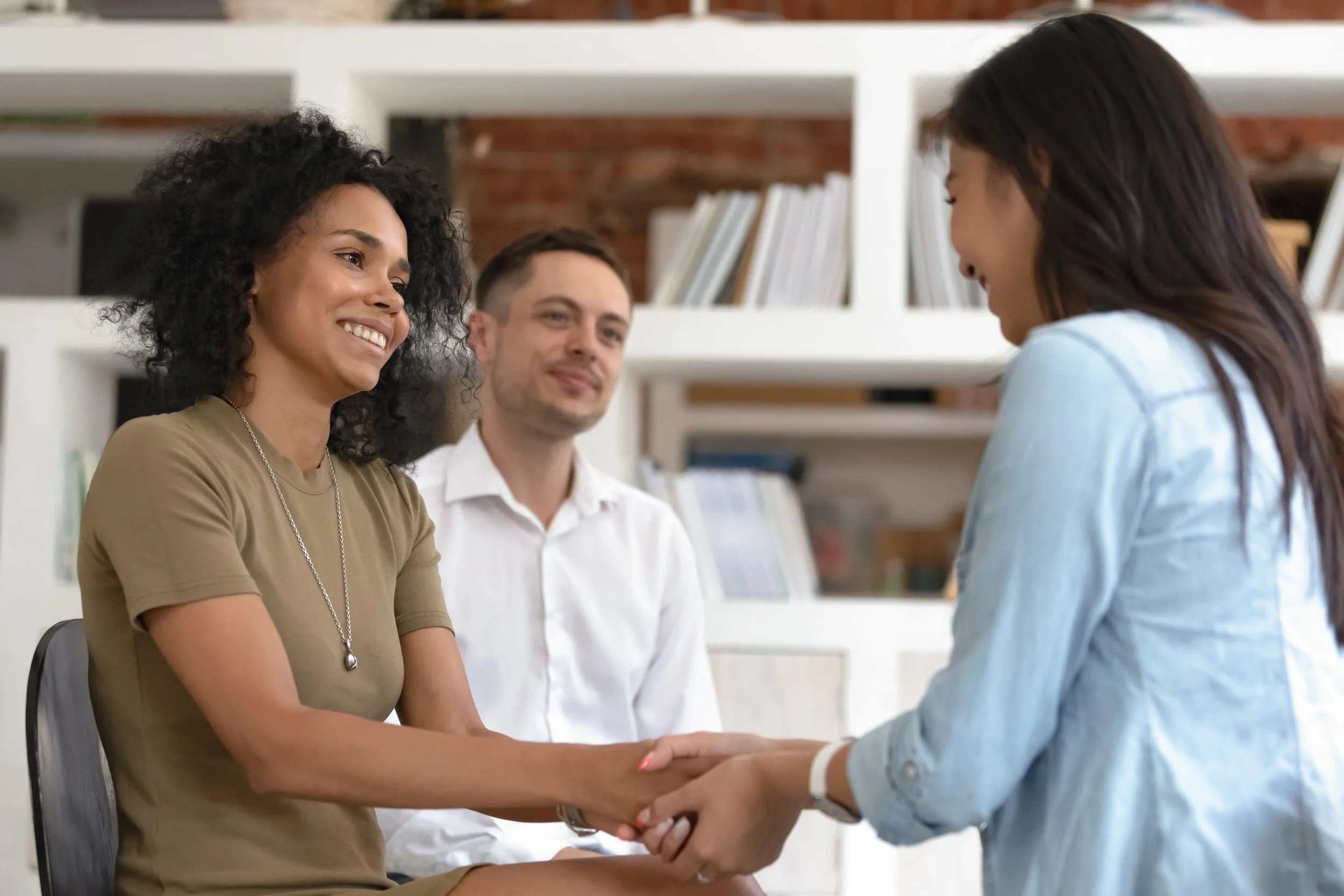 A woman smiling and receiving a handshake from another woman, with a man observing in the background, in a room with white shelves and books.