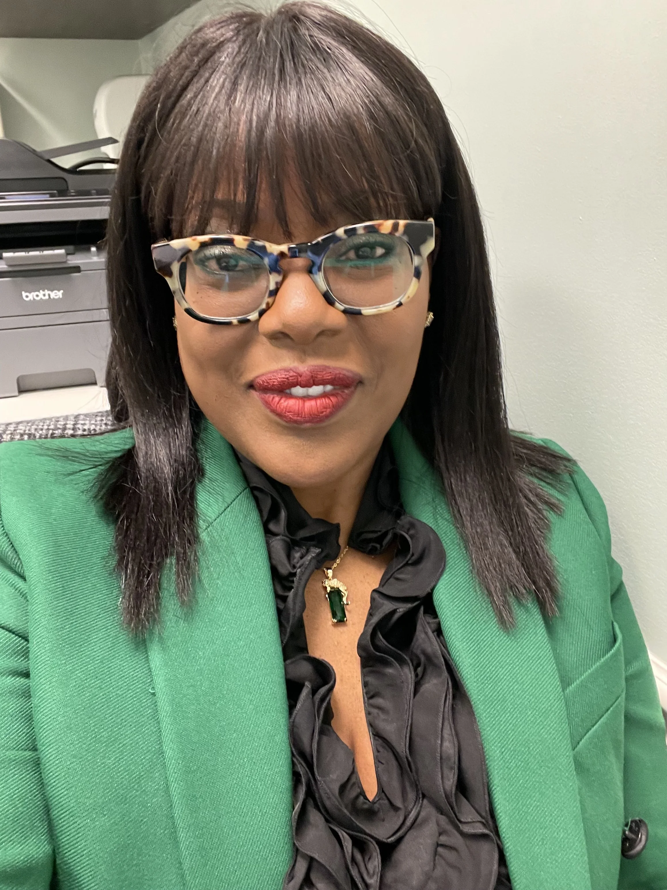 A woman wearing large leopard-print glasses, a green blazer, and a black ruffled top, with a necklace featuring a large square green pendant, sitting in an office environment with a gray printer in the background.