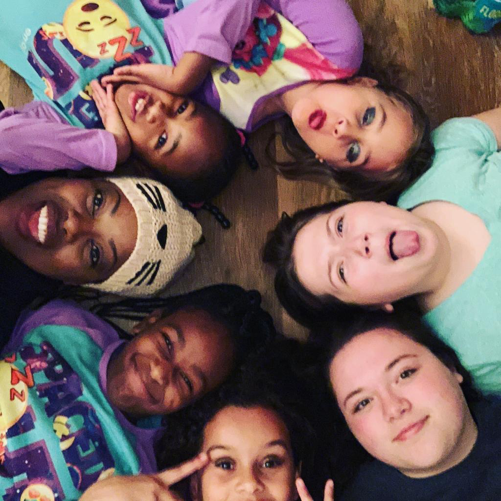 Group of young girls lying on the floor in a circle, making funny faces and smiling, with colorful pajamas and a wooden floor background.
