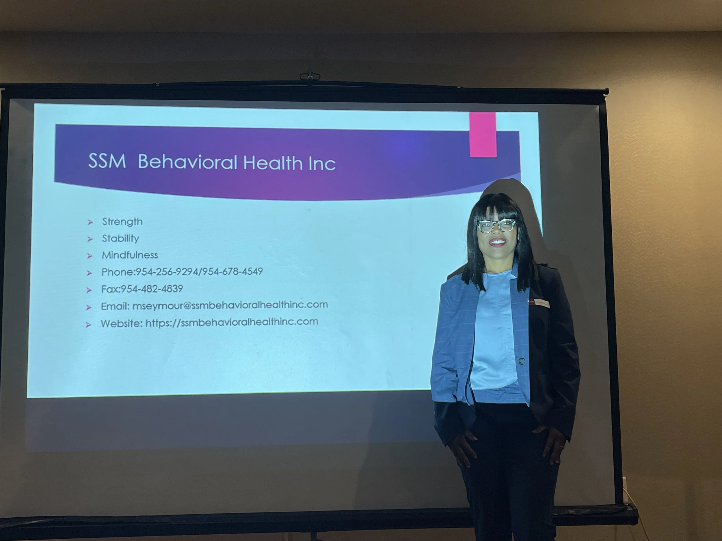 A woman in professional attire, wearing glasses, stands in front of a presentation slide that reads 'SSM Behavioral Health Inc' with contact information. The slide lists services such as strength, stability, mindfulness, with a phone number, fax, email, and website.