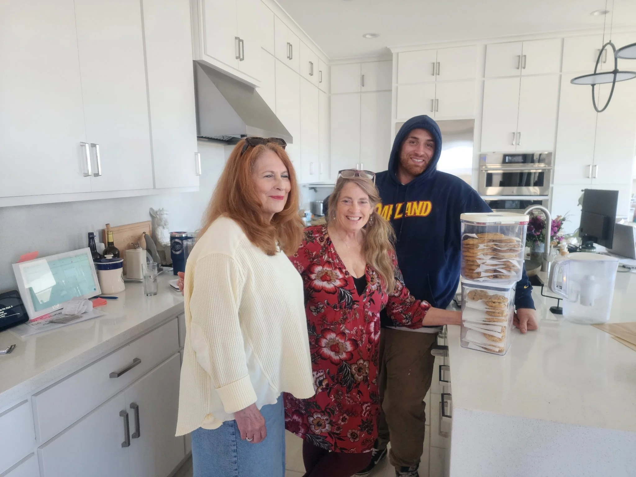 Three women and one man standing in a modern, white kitchen. The woman on the left has red hair and is wearing a cream sweater. The woman in the middle has long blonde hair and is wearing glasses and a red floral dress. The woman on the right has blonde hair and is dressed in a black shirt. The man on the far right has a beard, is wearing a hoodie, and is smiling. There are snacks and beverages on the counter, and the kitchen has white cabinets, a stainless steel oven, and a decorative light fixture hanging from the ceiling.