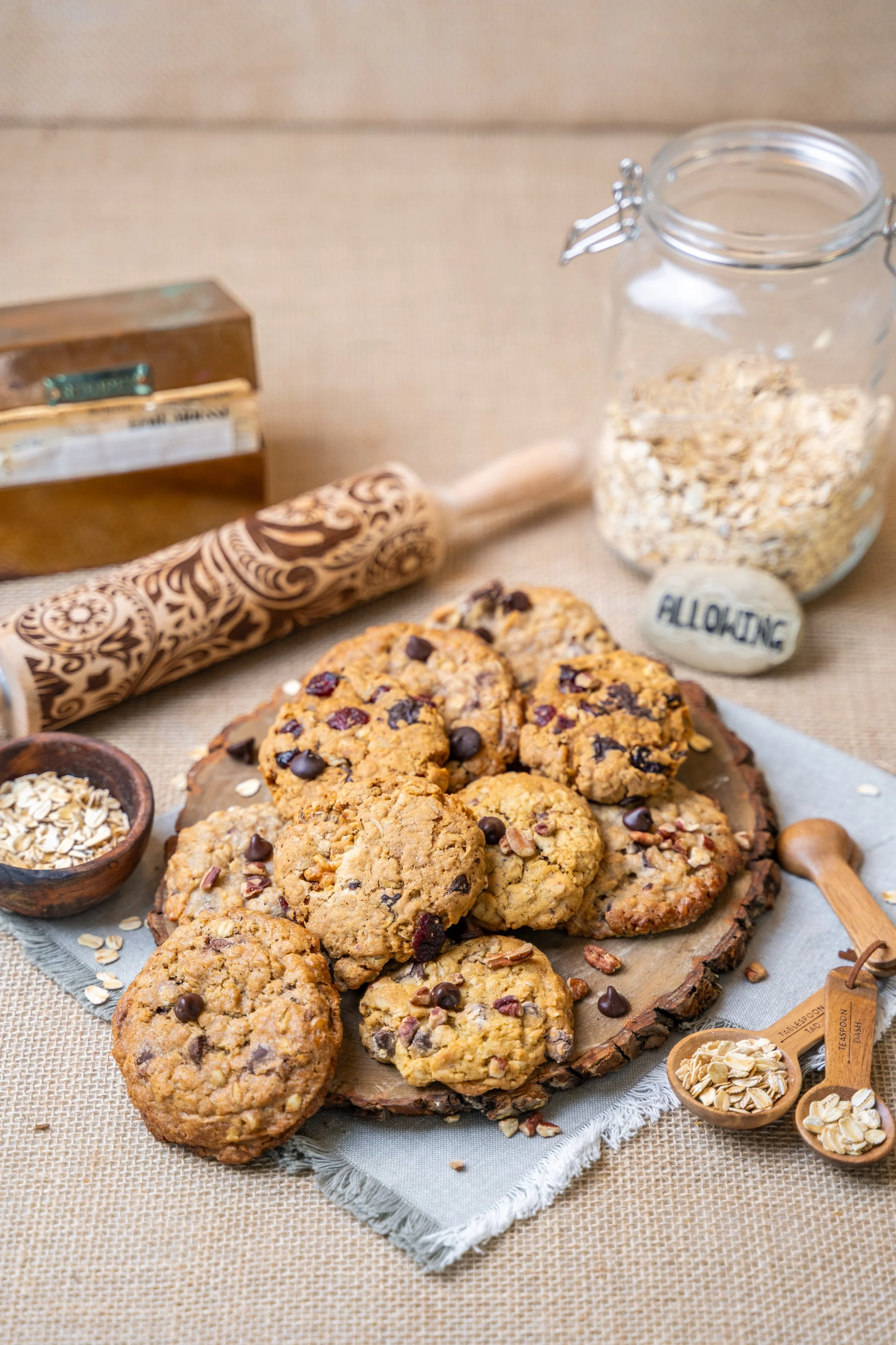 Oatmeal cookies with chocolate chips and chopped nuts on a wooden serving board, with small bowls of oats and chopped nuts, and a jar of oats in the background.