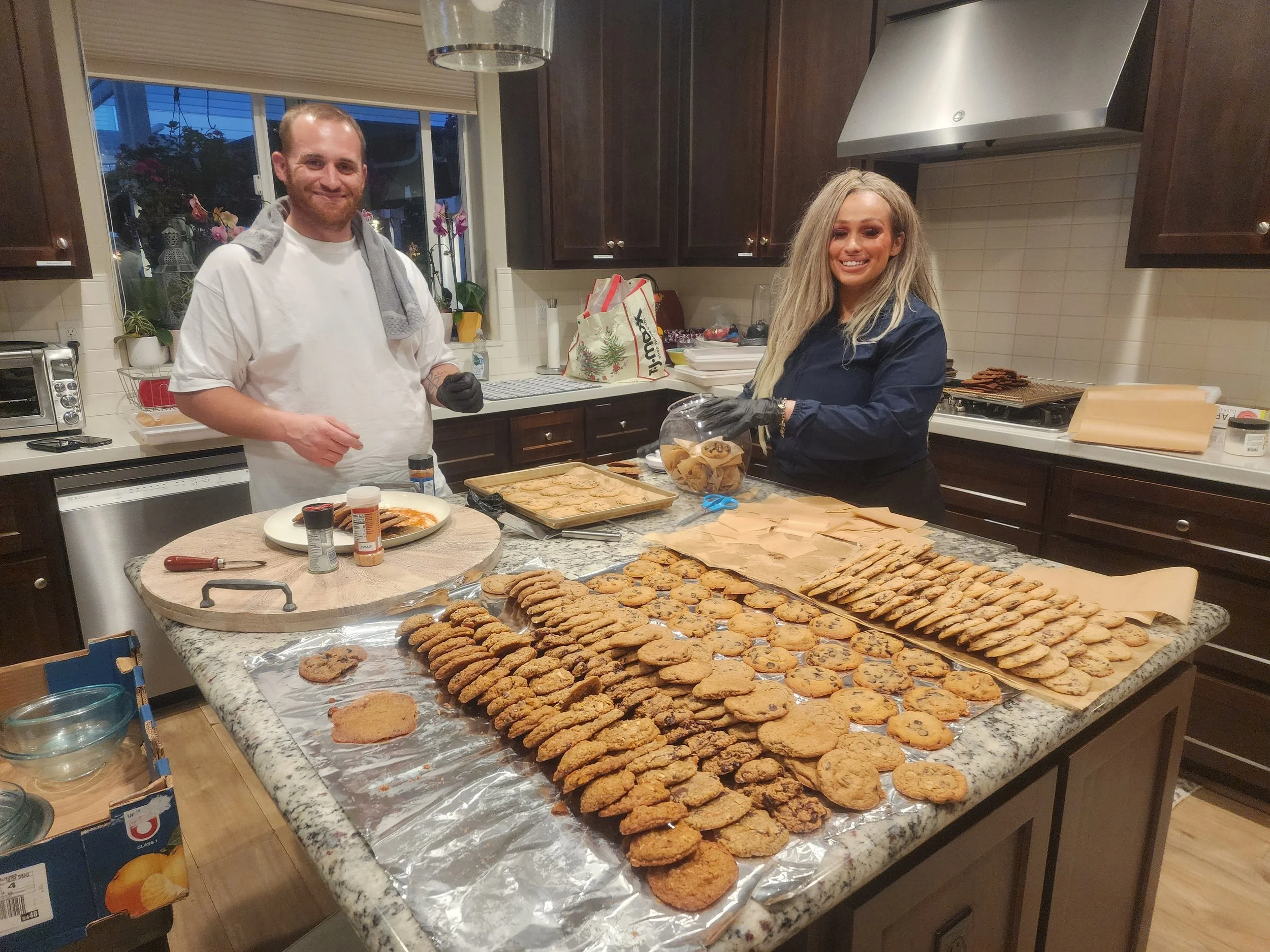 A man with a short beard and wearing a white shirt and black gloves, and a woman with long blonde hair and wearing a dark jacket, prepare and display an extensive spread of cookies on a kitchen counter. The cookies are arranged in rows on parchment paper, with some in clear plastic containers. The kitchen features dark wooden cabinets, a window with potted plants, and various baking supplies and gifts on the counter.