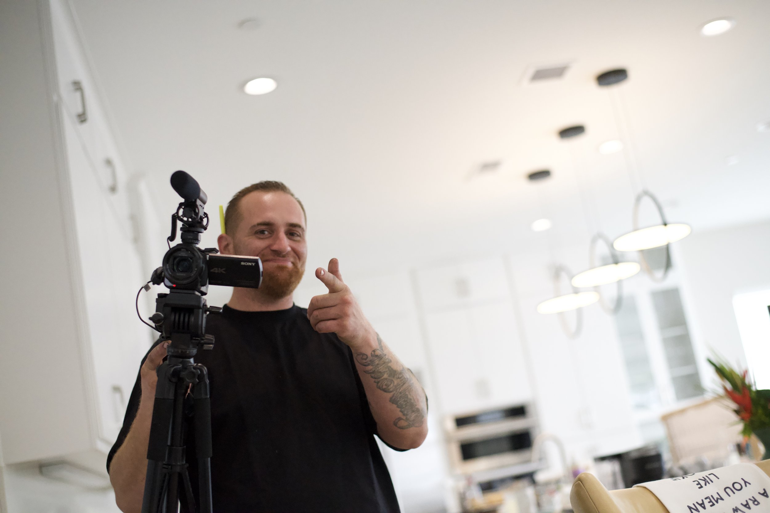 A man with a beard and tattoos posing for a selfie, holding a video camera on a tripod, in a room with white ceilings and round ceiling lights.