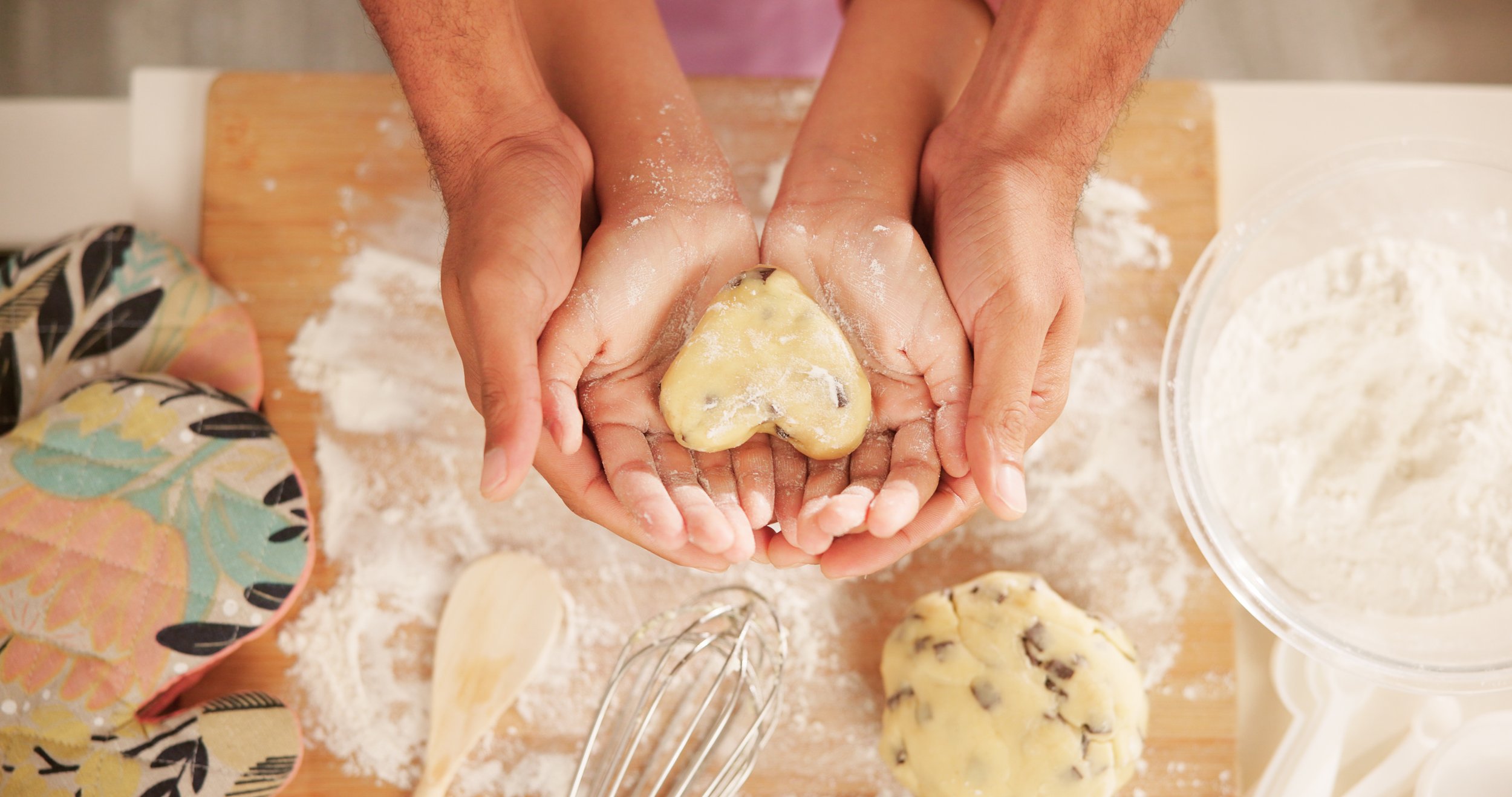 Hands pressing down on cookie dough with chocolate chips, surrounded by baking tools and ingredients on a floured surface.