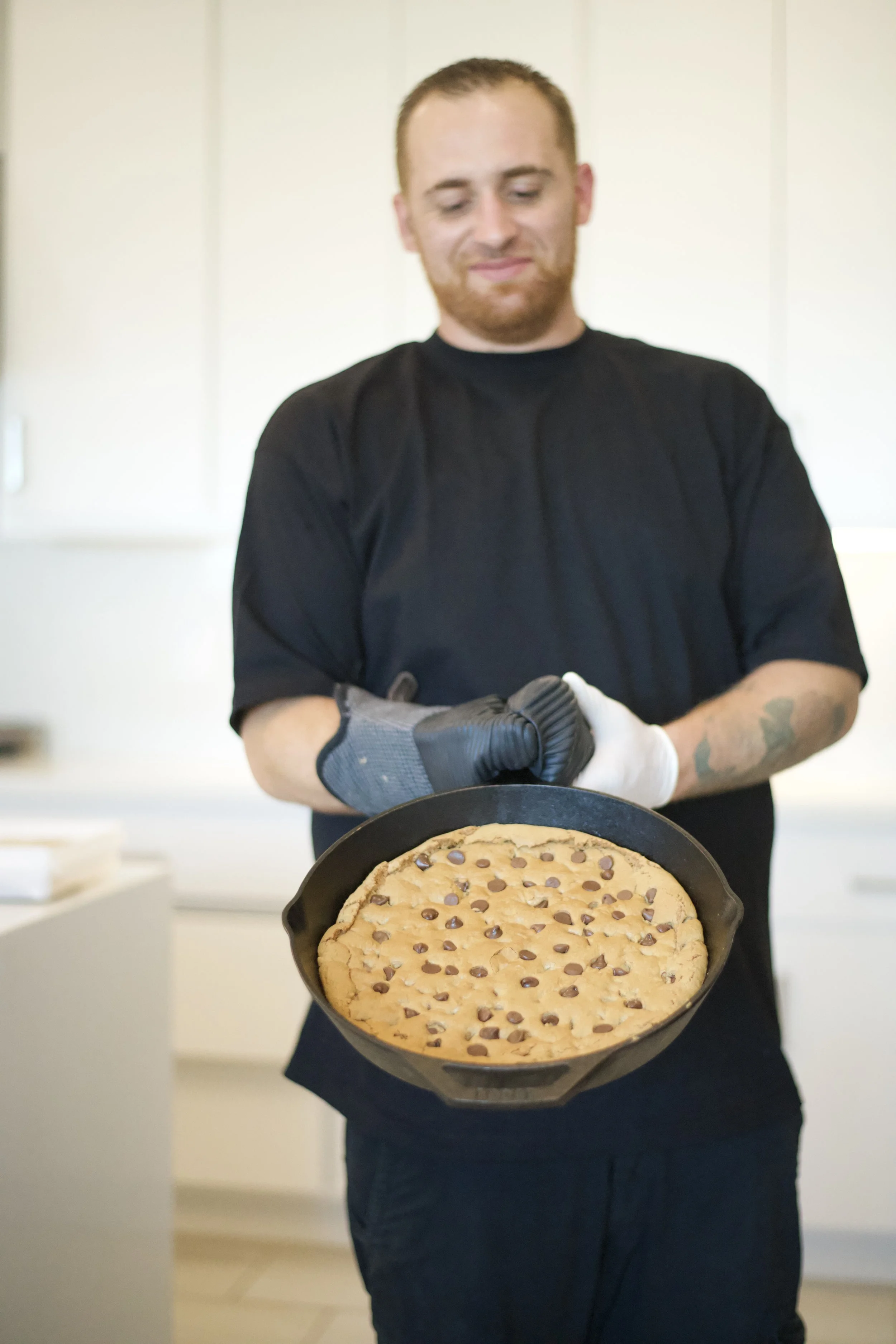 A man with tattoos on his arm, wearing a black shirt, is holding a freshly baked chocolate chip cookie cake in a cast iron skillet, standing in a bright kitchen.