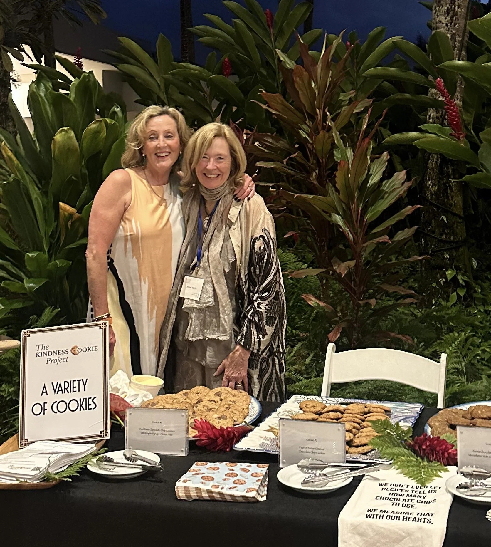 Two women standing and smiling behind a table filled with various cookies at a charity event. There are decorative plants and signs discussing cookies and gratitude.