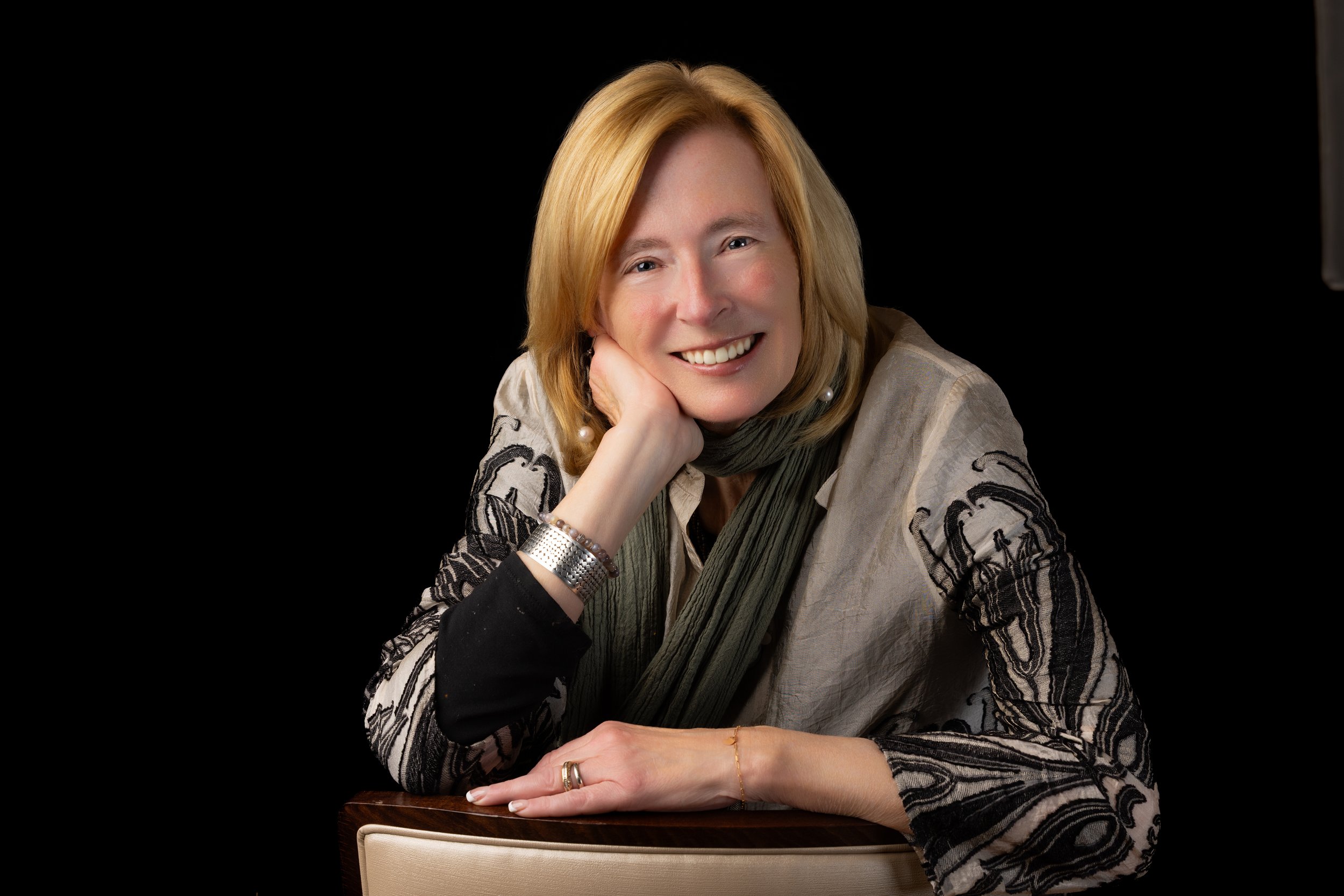 A woman with blonde hair, smiling, resting her head on her left hand, sitting at a table with a black background.